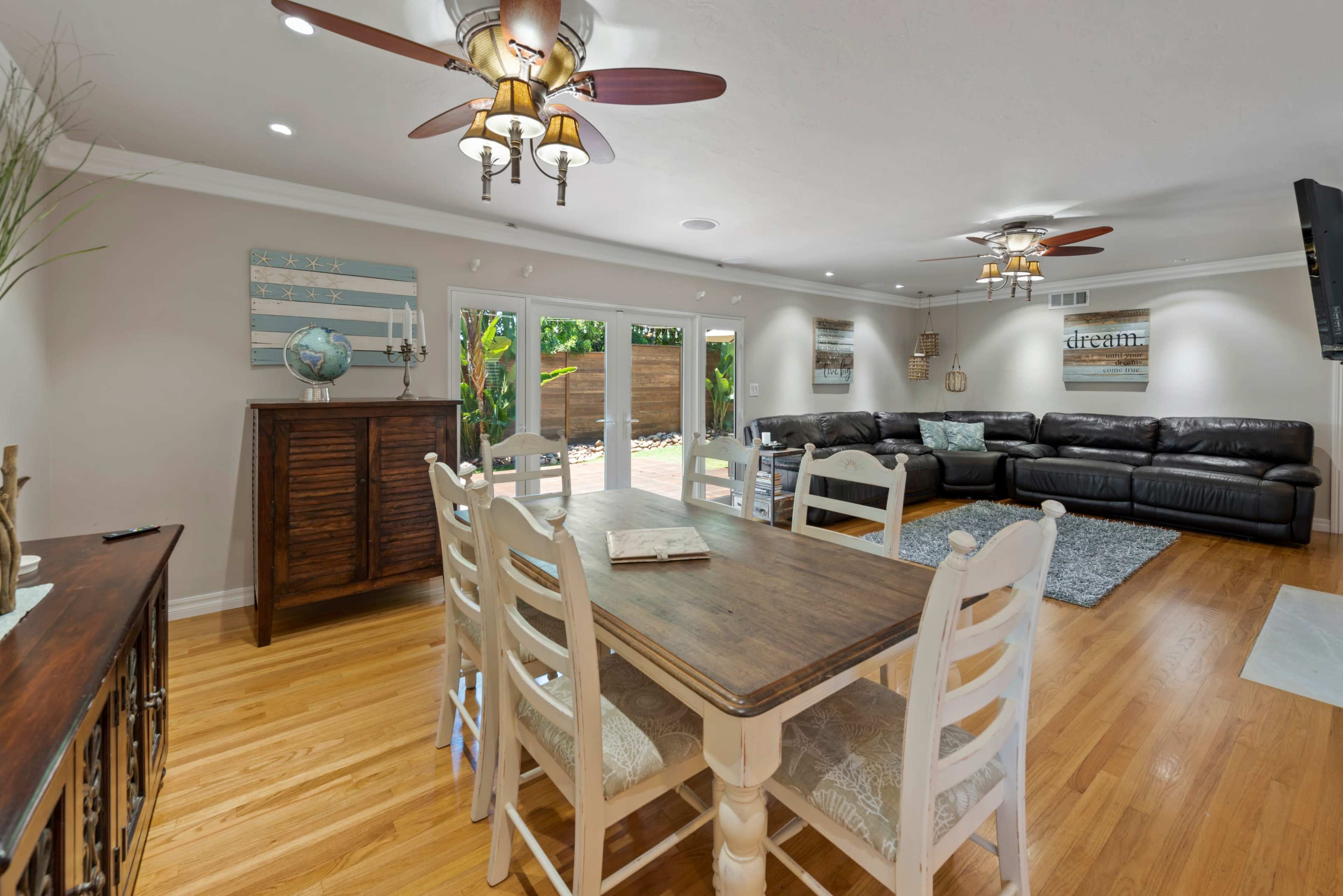 A spacious living and dining area features a wooden table surrounded by white chairs, a leather sectional sofa, and large sliding glass doors leading to an outdoor space.