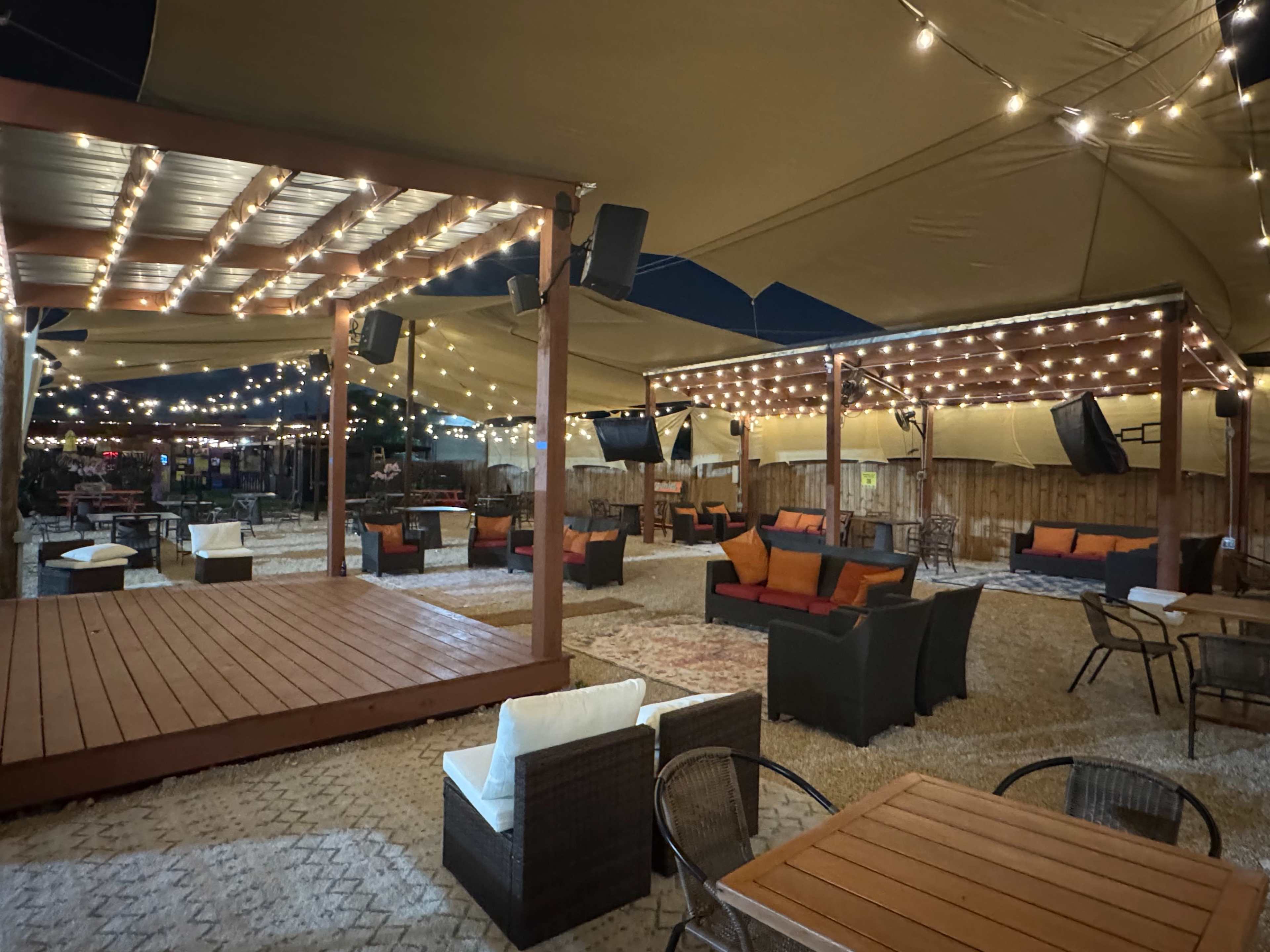 The image shows an outdoor seating area with string lights overhead, featuring multiple tables and lounge seating arranged under a large canopy.