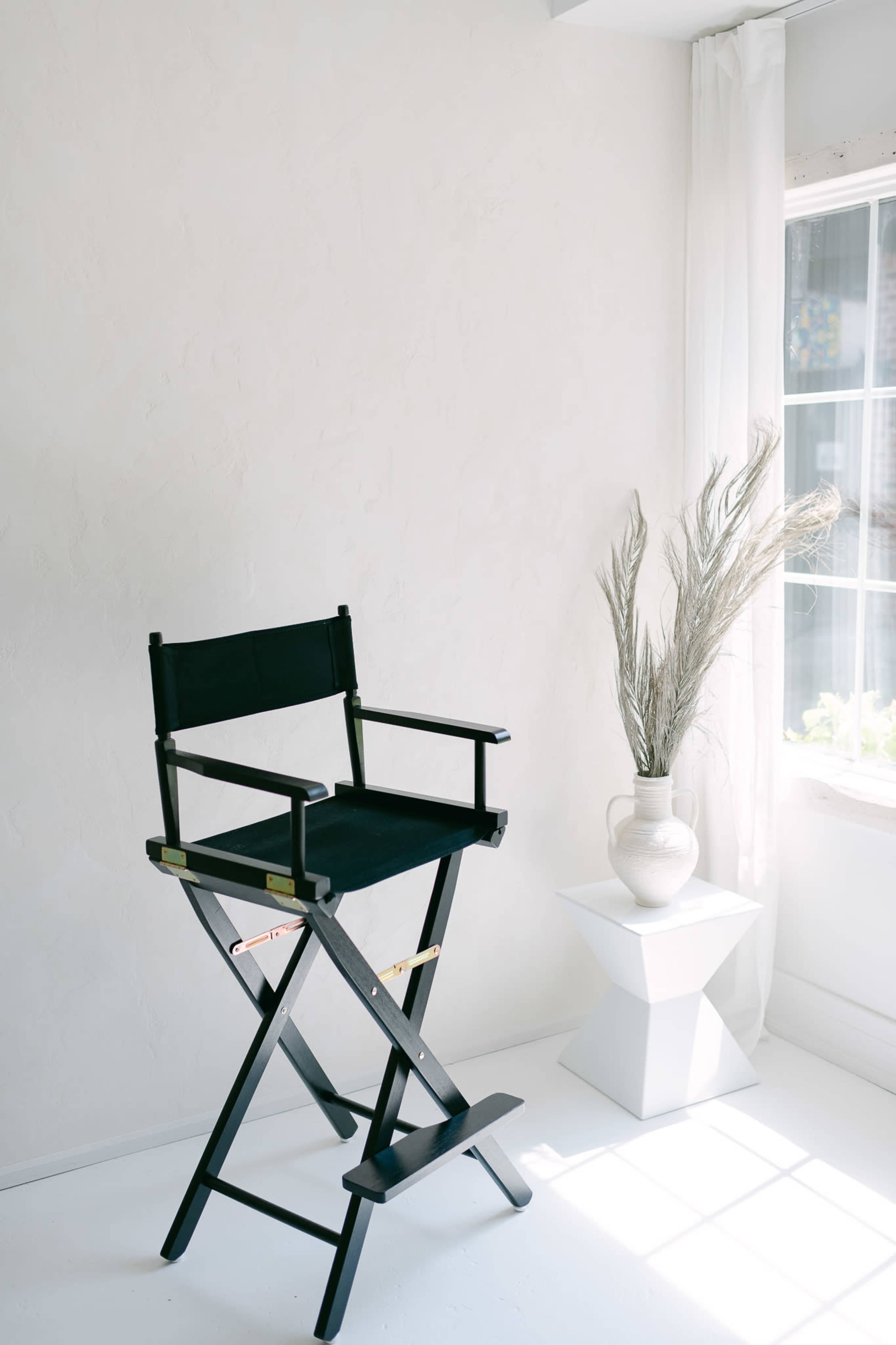 A black director's chair stands next to a white pedestal table and a vase with dried grass in a bright, minimalist room.
