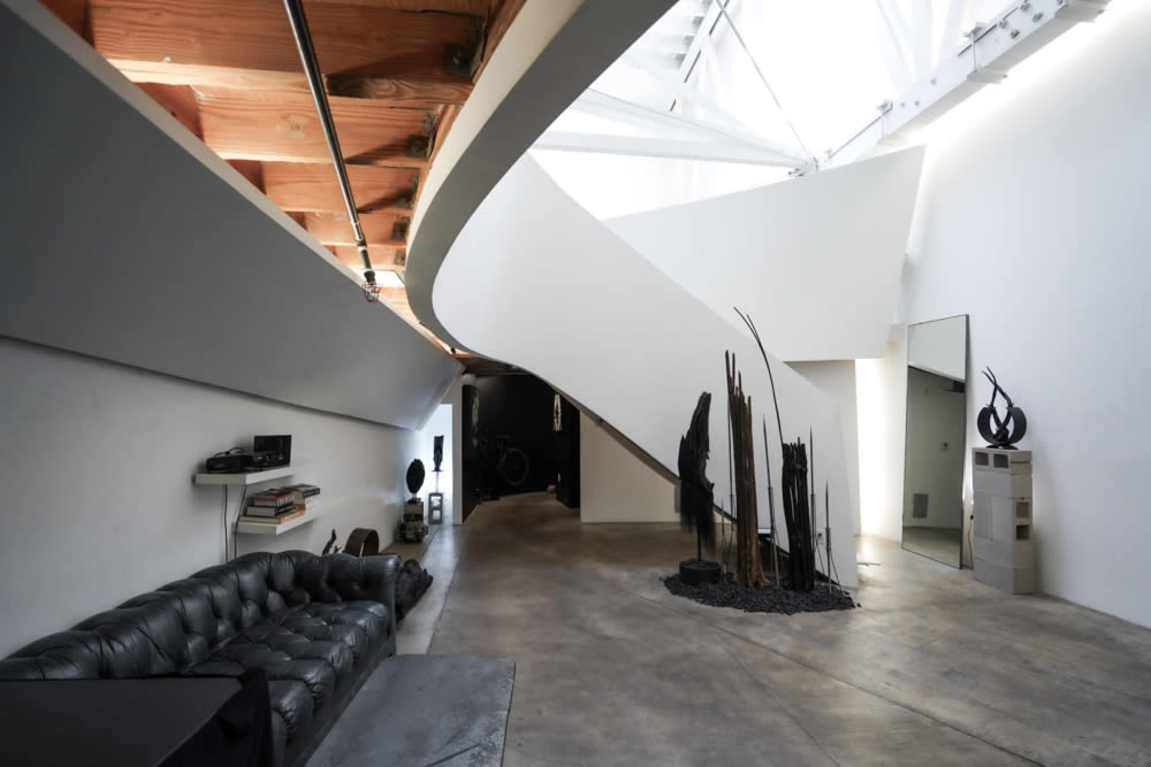 A modern interior space featuring a curved staircase, a black leather couch, and abstract sculptures arranged on the floor.