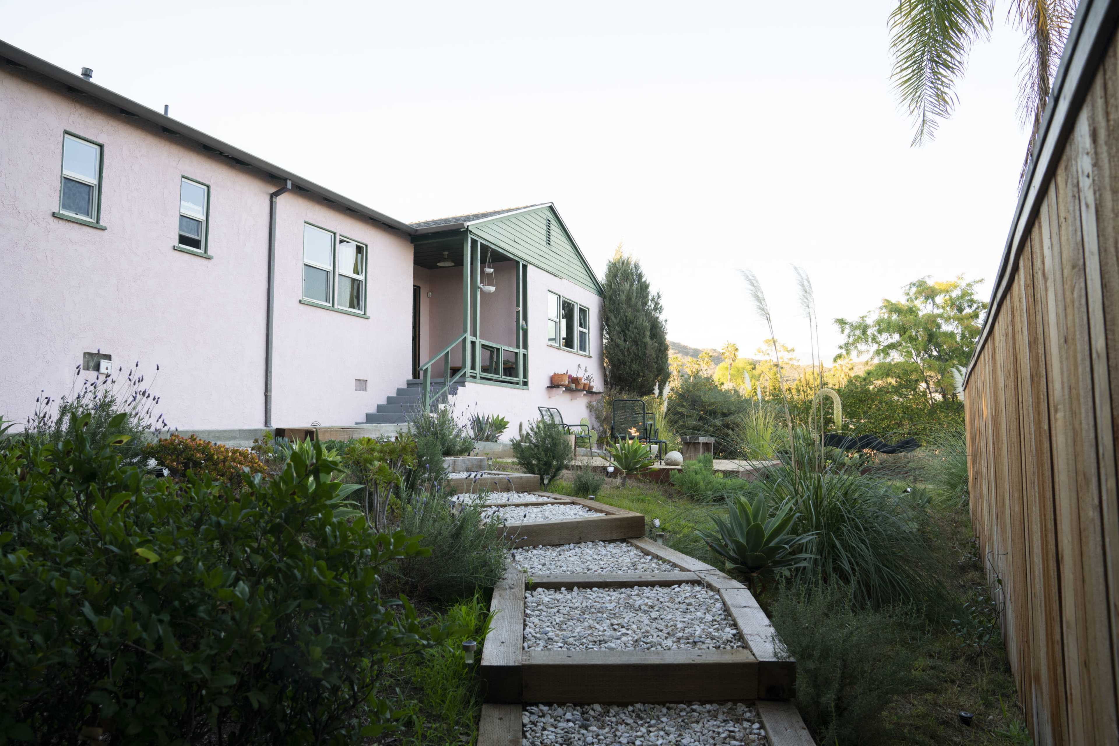 A pathway with gravel and wooden edging leads to a light pink house with green trim and a porch, surrounded by various plants and trees.
