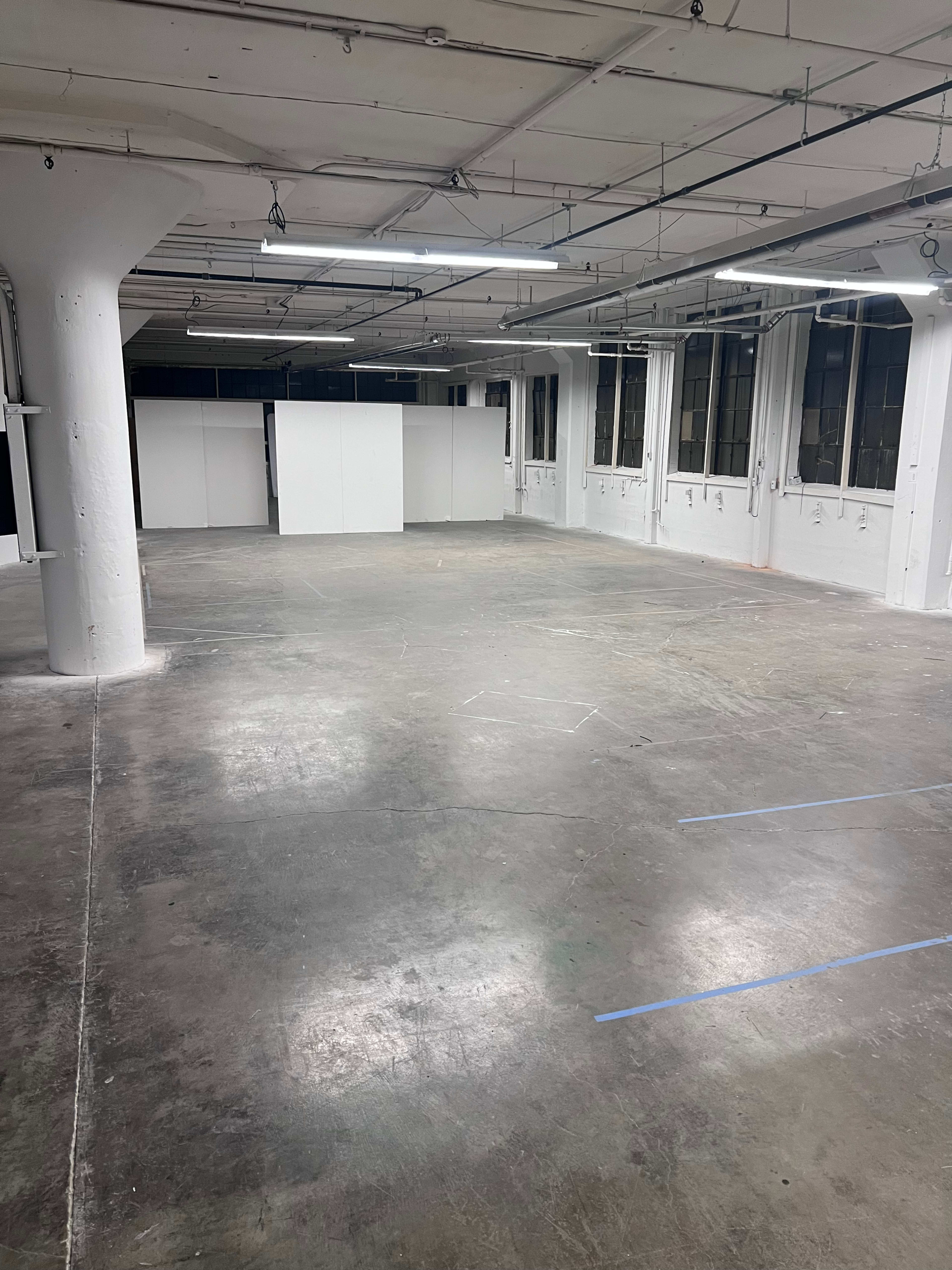 The image shows a spacious, empty interior room with polished concrete floors and large windows, featuring white walls and a few partitions in the background.