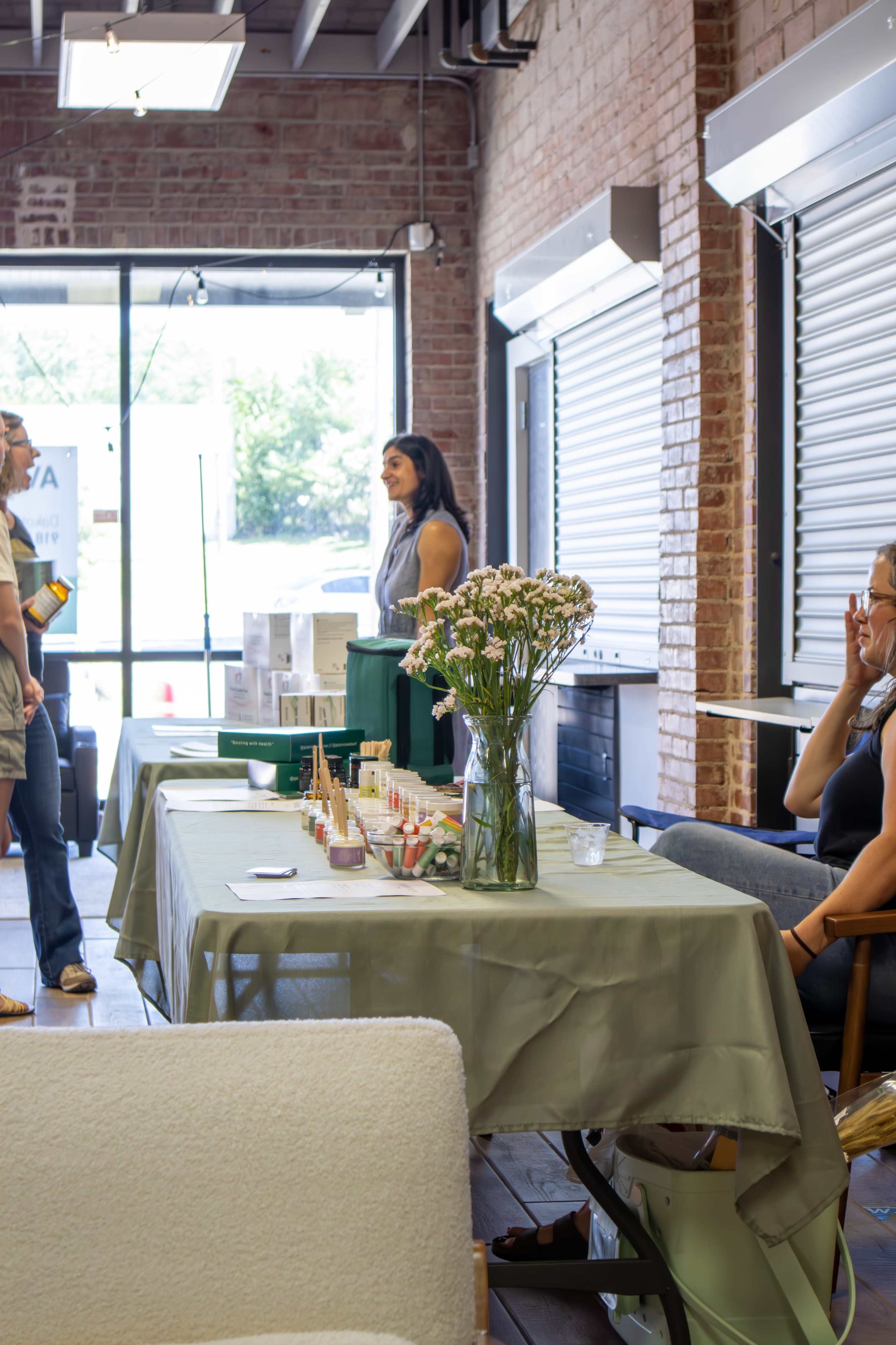 A woman stands behind a table displaying cosmetic products, while two other individuals engage in conversation in a well-lit, brick-walled space.
