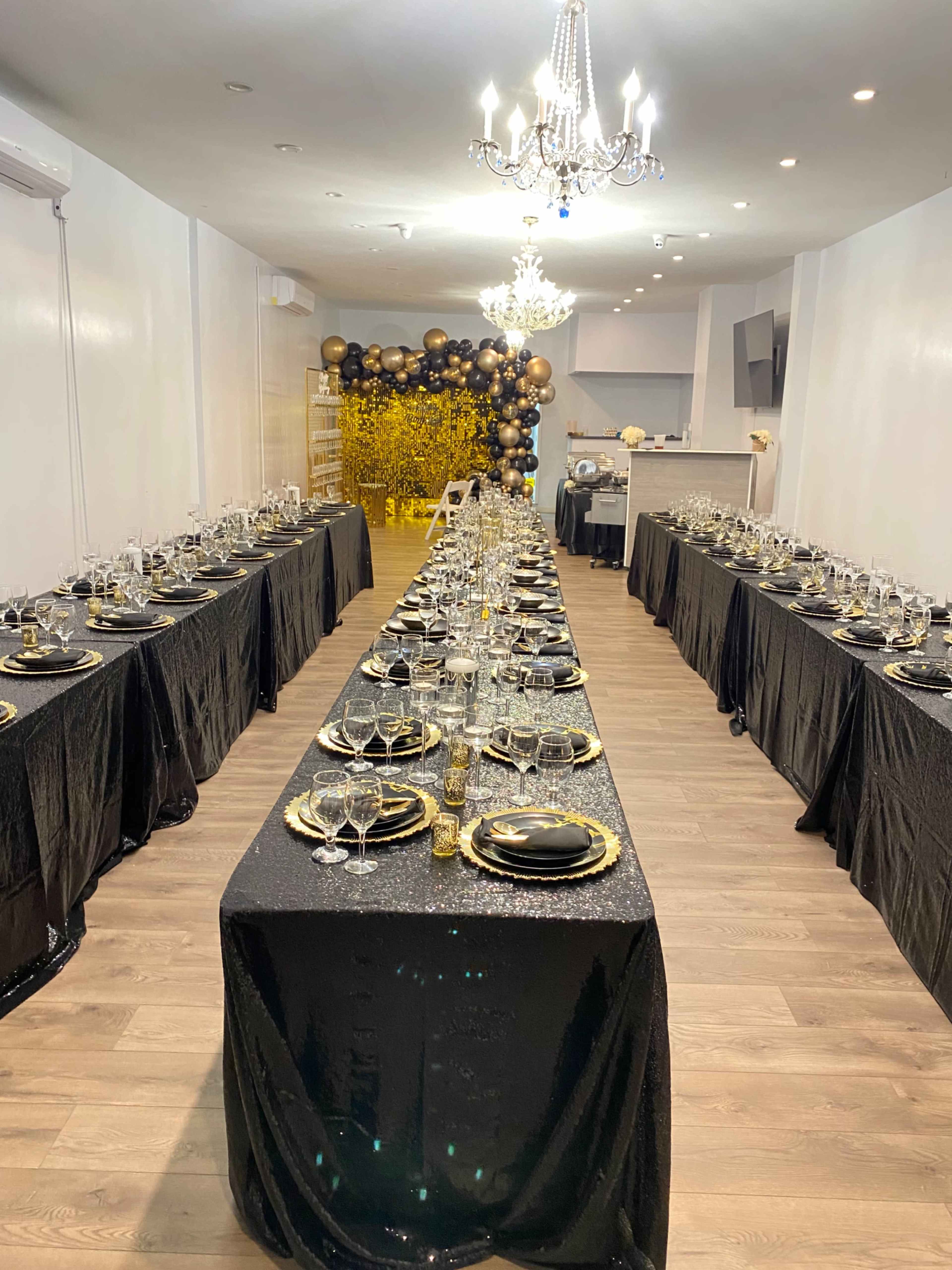 A large dining setup with black tables covered in sequined cloths, elegantly arranged with black plates, crystal glasses, and a gold-themed backdrop featuring balloons and decorations.