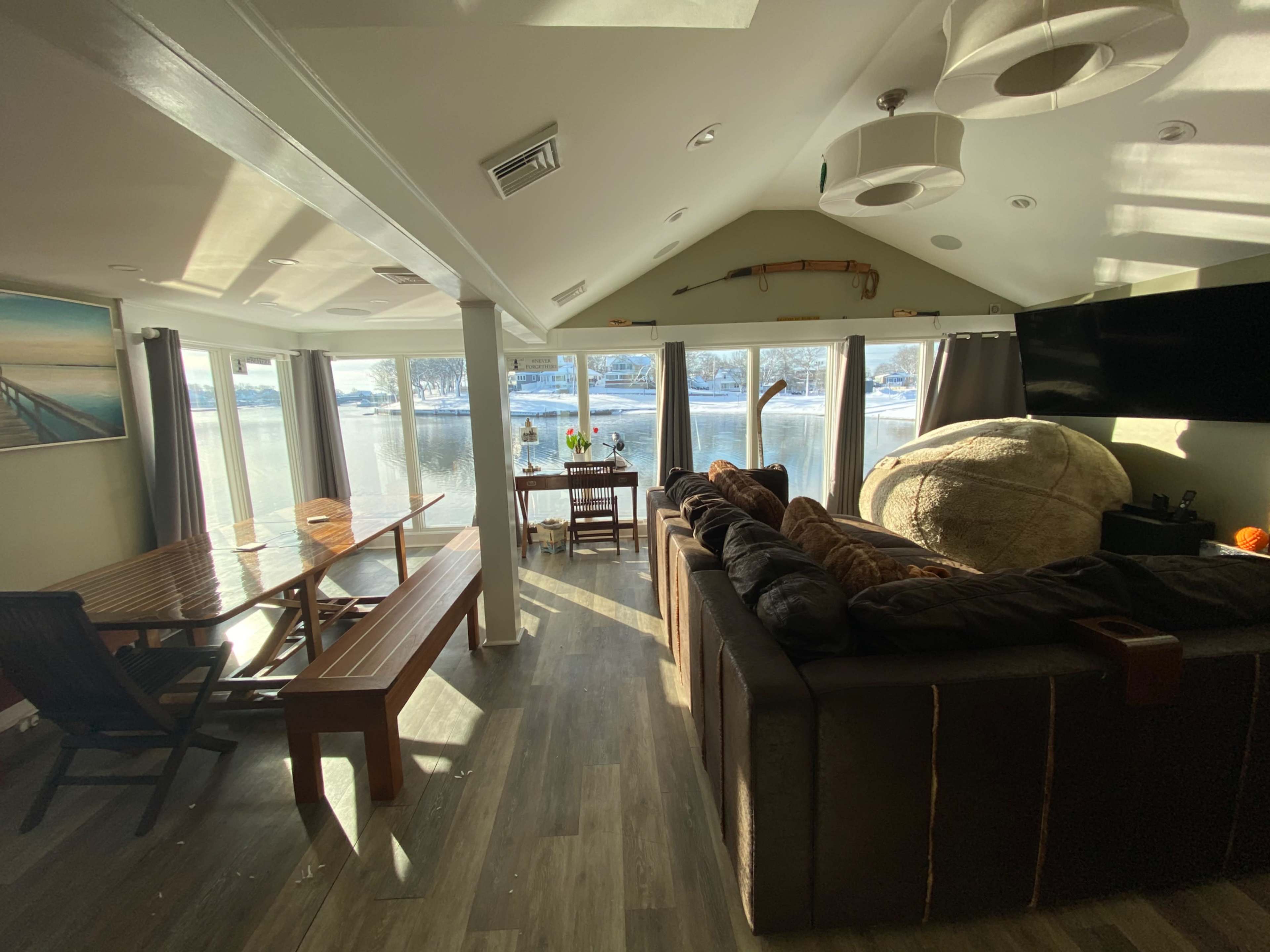 The image shows a cozy living room with large windows overlooking a lake, featuring a sectional sofa, a wooden dining table, and a comfortable chair.
