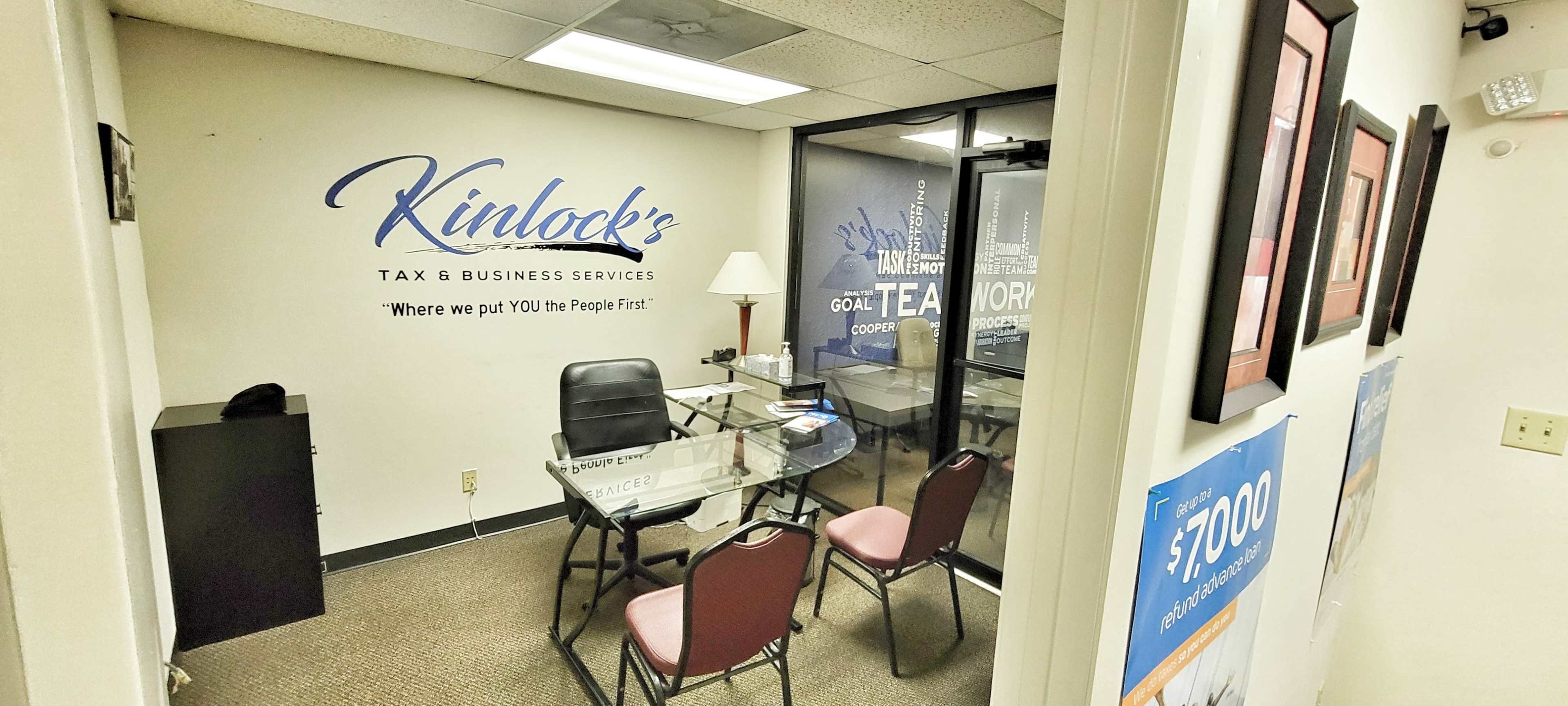 The image shows a small office space with a glass wall, a desk with a chair, and promotional materials on the walls.