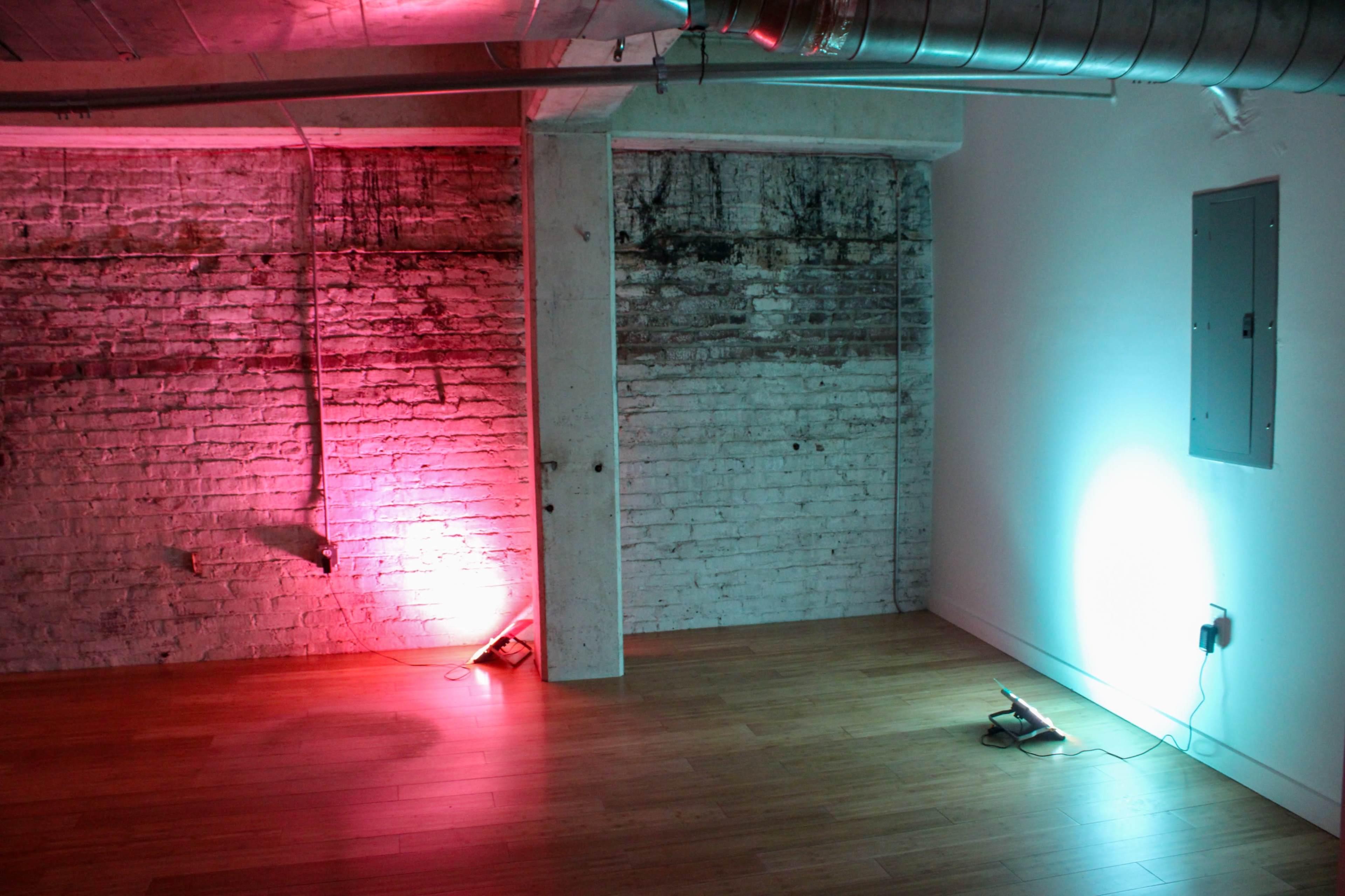 The image shows an empty room with wooden flooring, featuring two contrasting light sources illuminating the walls with pink and blue hues.