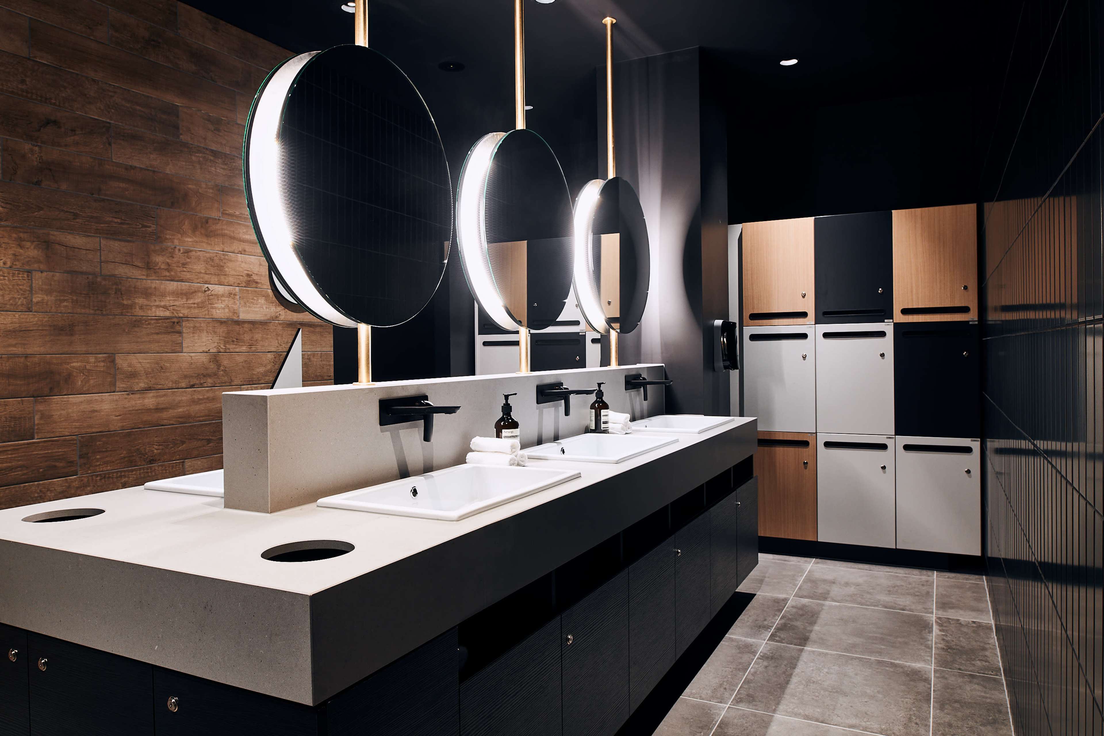 The image shows a modern bathroom with three sinks under circular mirrors, illuminated by overhead lights, accompanied by wooden and metal lockers along the wall.