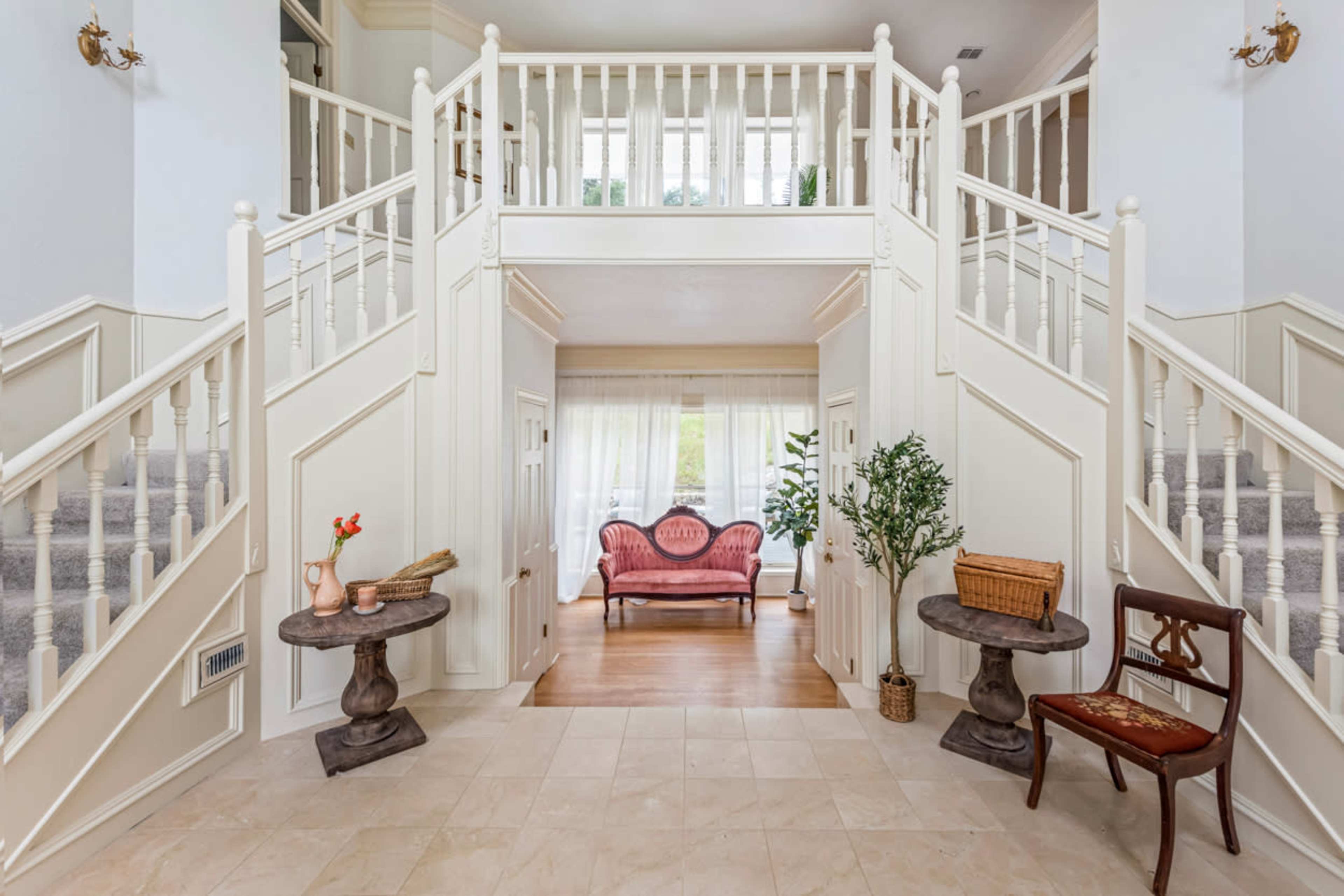 The image shows a spacious foyer with a grand staircase, two small round tables, a pink settee, and a single wooden chair.