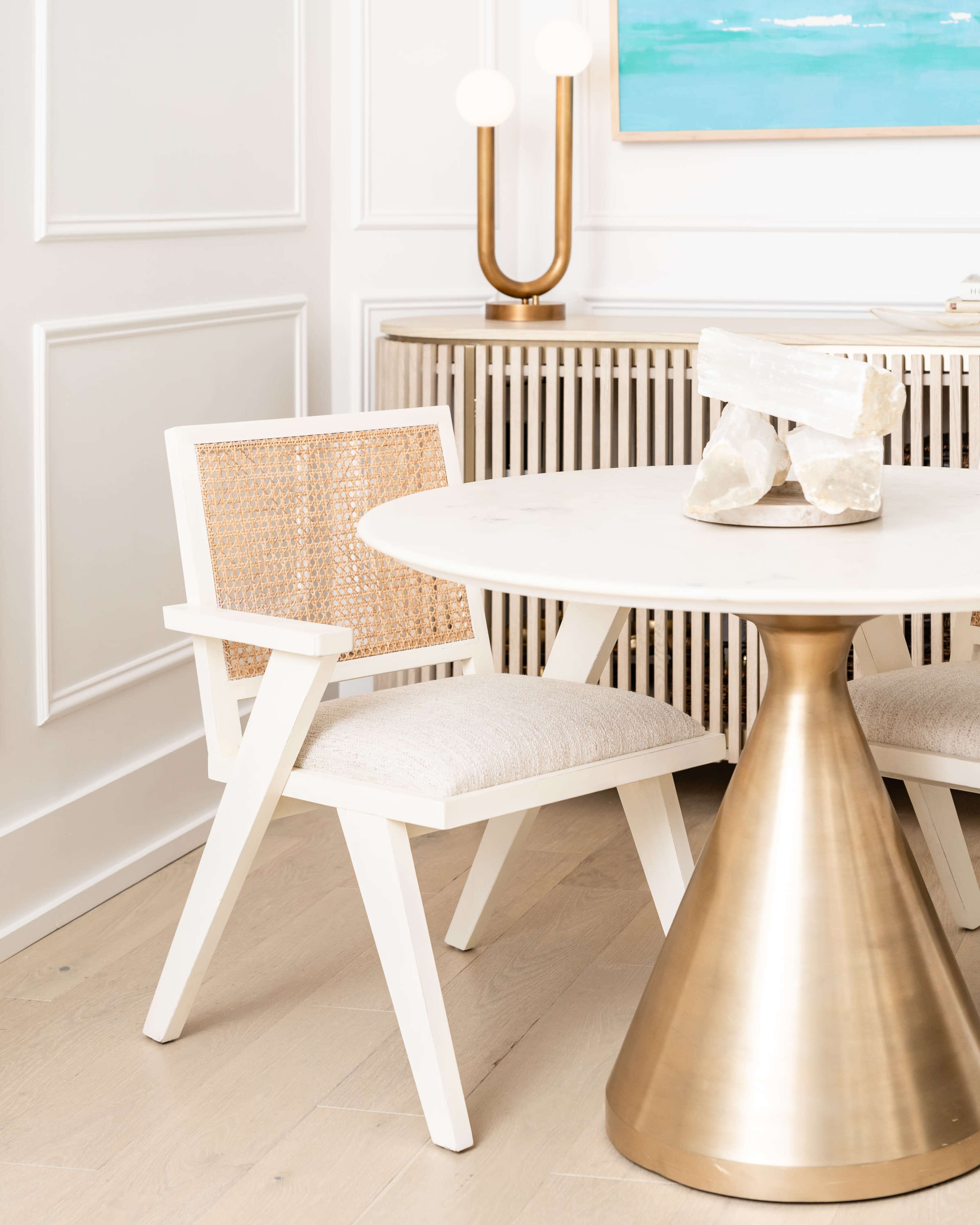 A modern dining area features a round table with a brass base, accompanied by a white chair with a woven back and light-colored upholstery.
