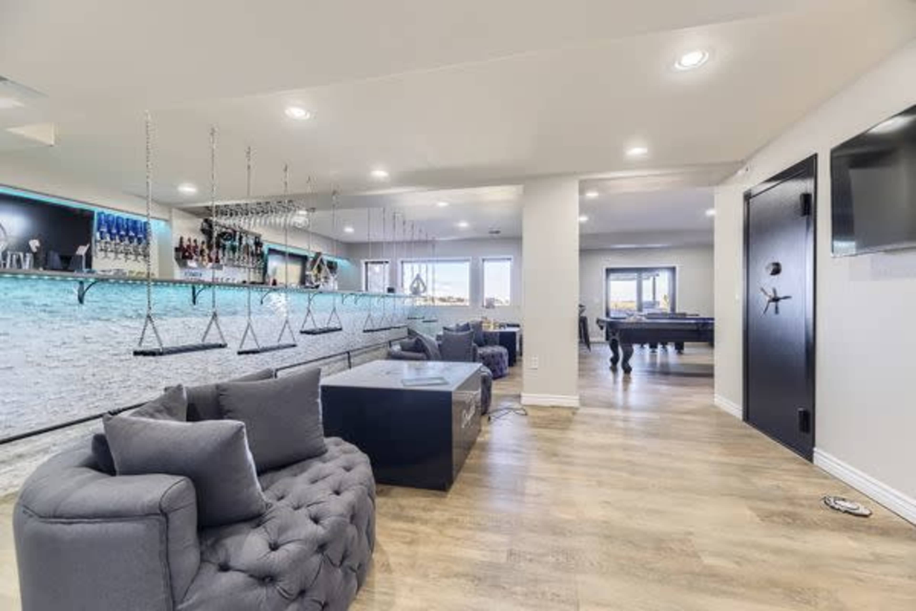 The image shows a modern indoor lounge area featuring a bar with hanging glassware, a sectional sofa, a dark coffee table, and a pool table in the background.