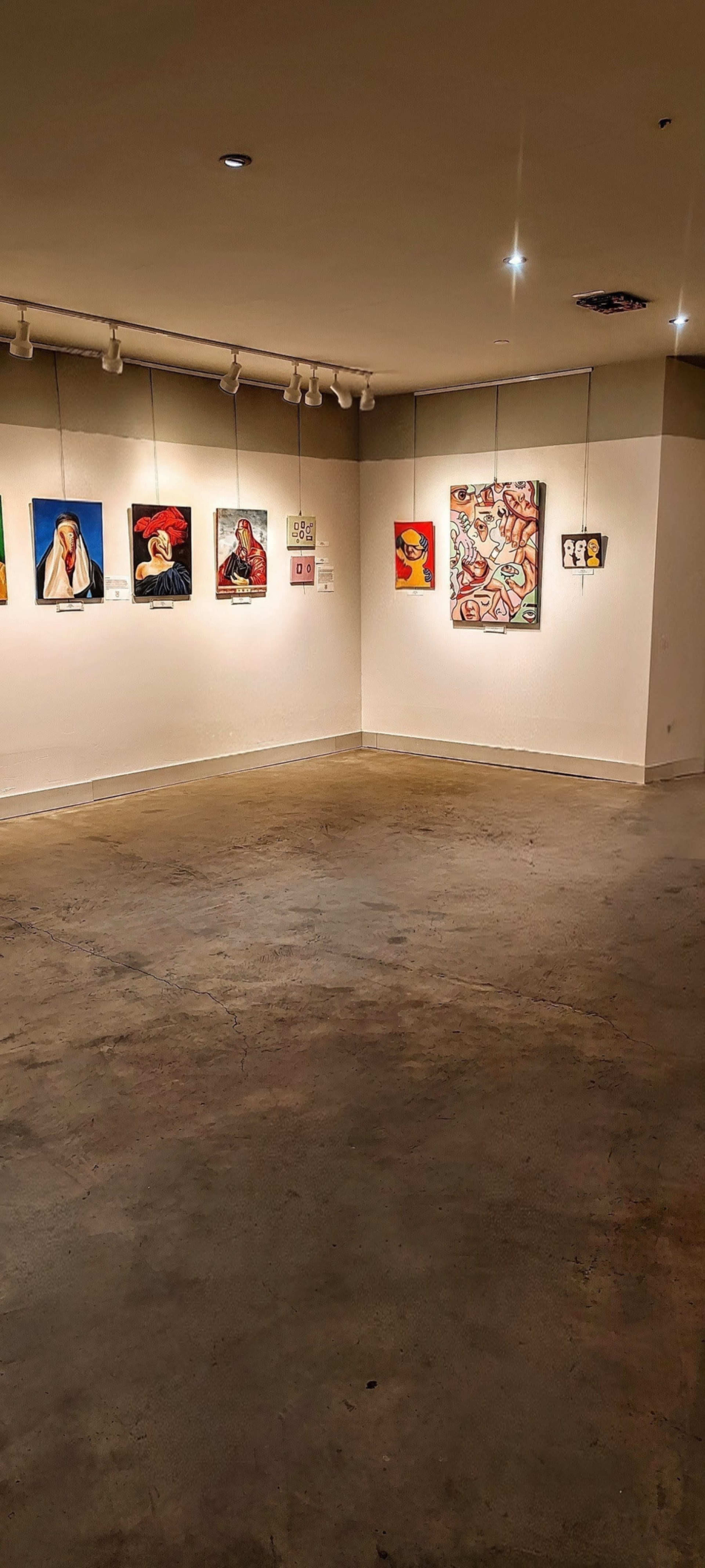 The image depicts a sparse art gallery with several paintings displayed on the walls and a concrete floor.
