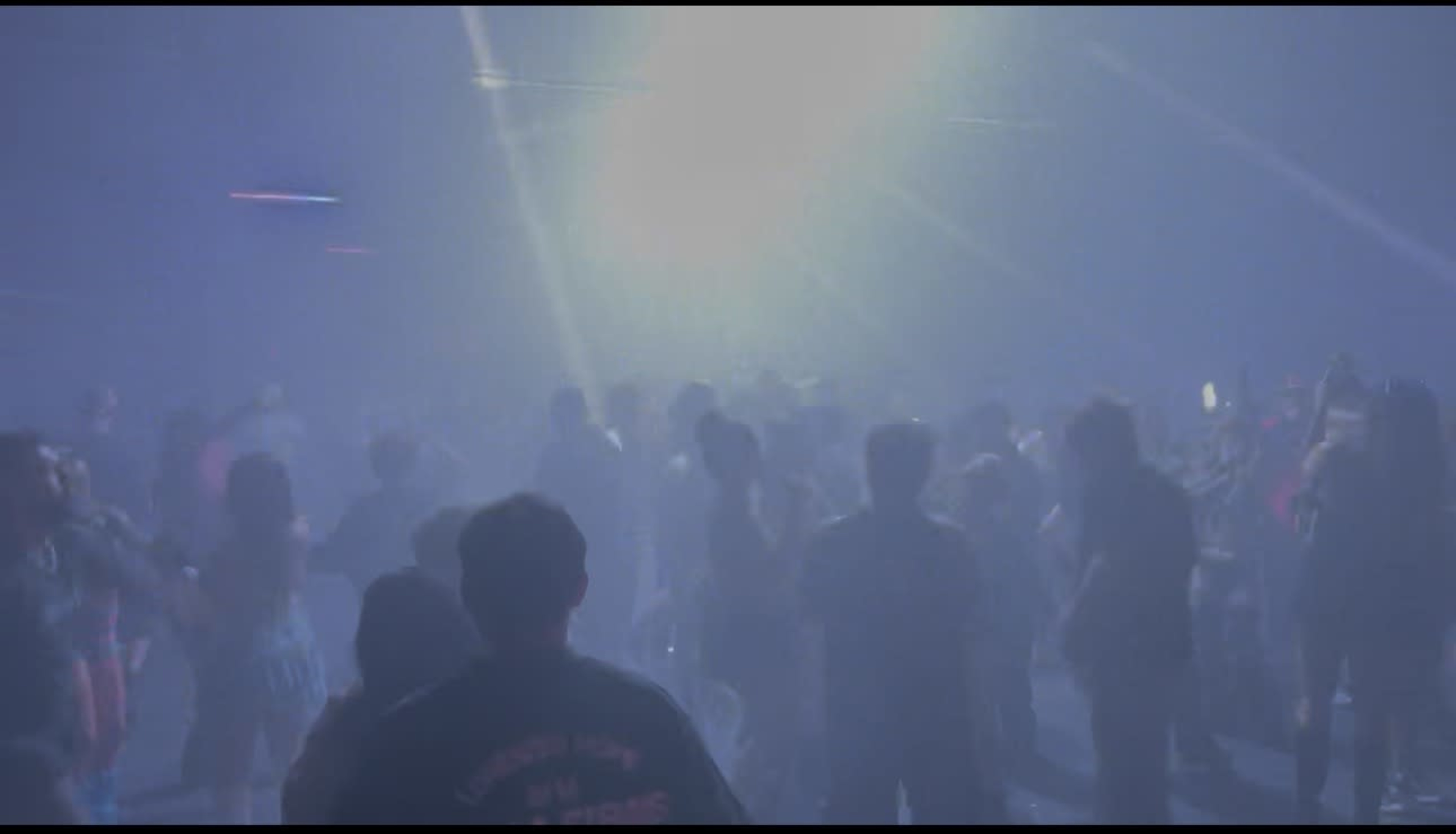 A crowd of people dances in a dimly lit venue filled with fog and colorful lights.