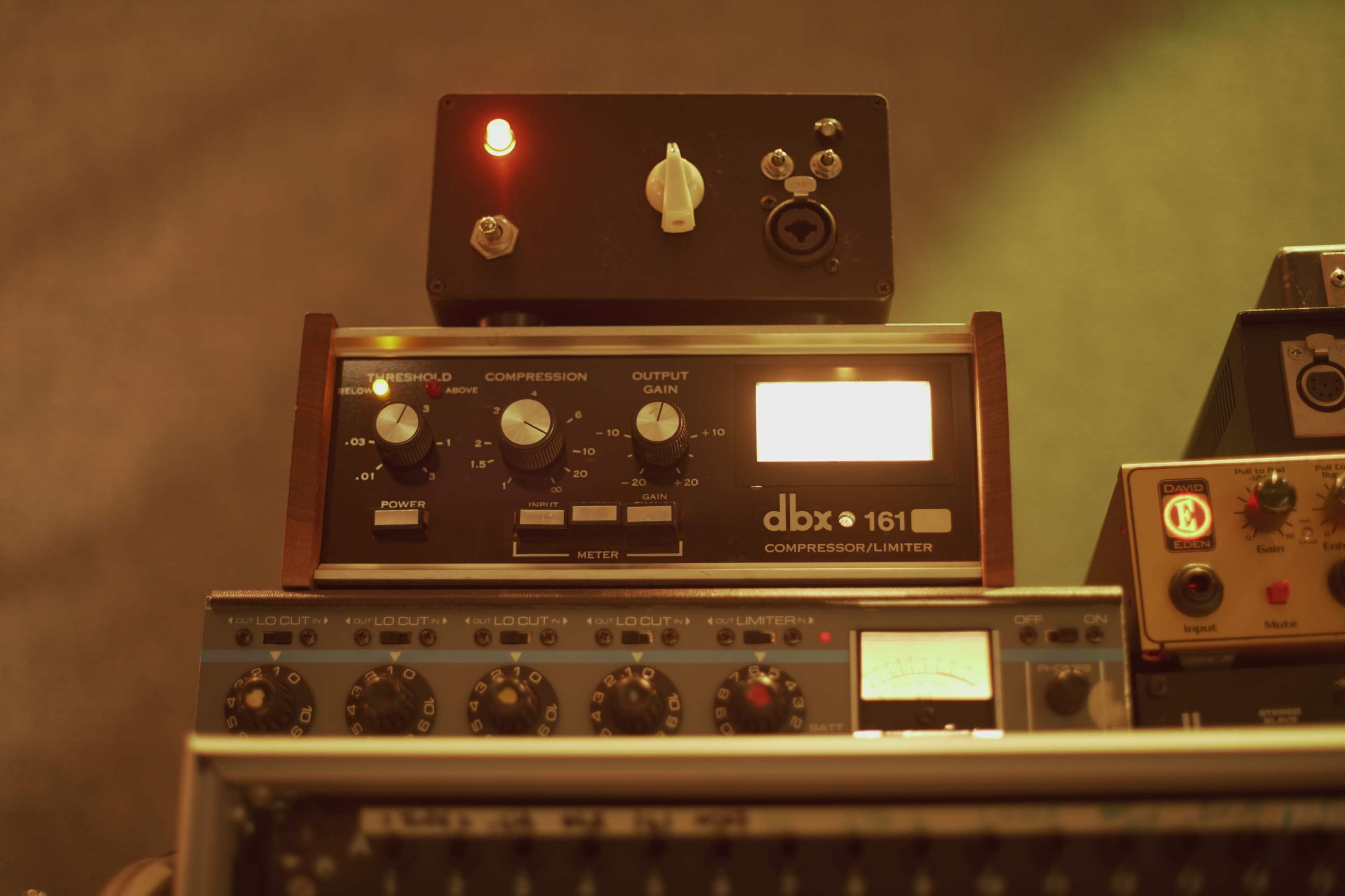 The image shows a dbx 161 compressor/limiter unit with various knobs, a meter, and an illuminated power indicator displayed on a rack along with other audio equipment.