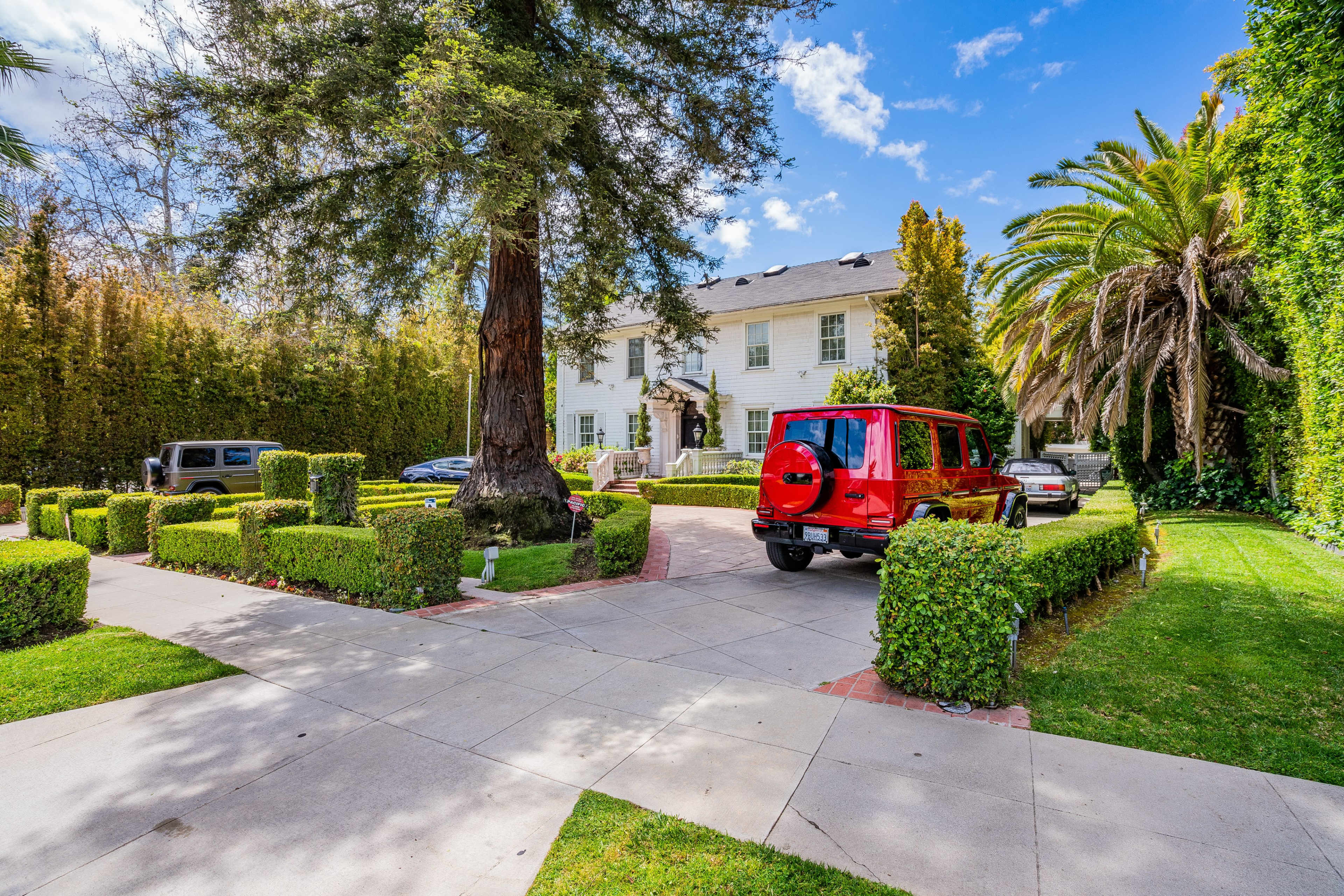 A red SUV is parked in the driveway of a large white house surrounded by well-manicured hedges and palm trees.