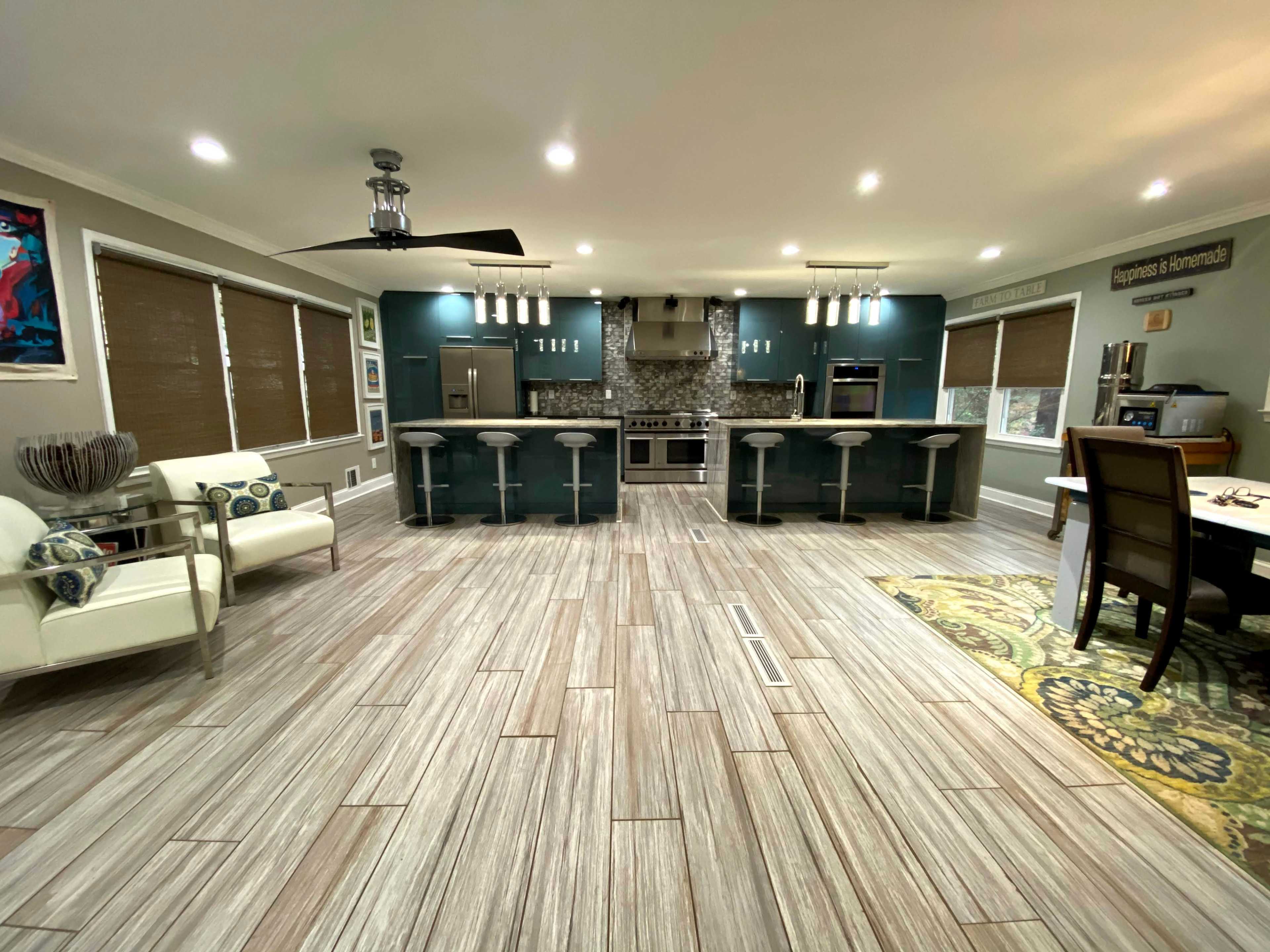 The image shows a modern kitchen and dining area featuring a central island with bar stools, large windows, and a combination of tiled and wooden flooring.