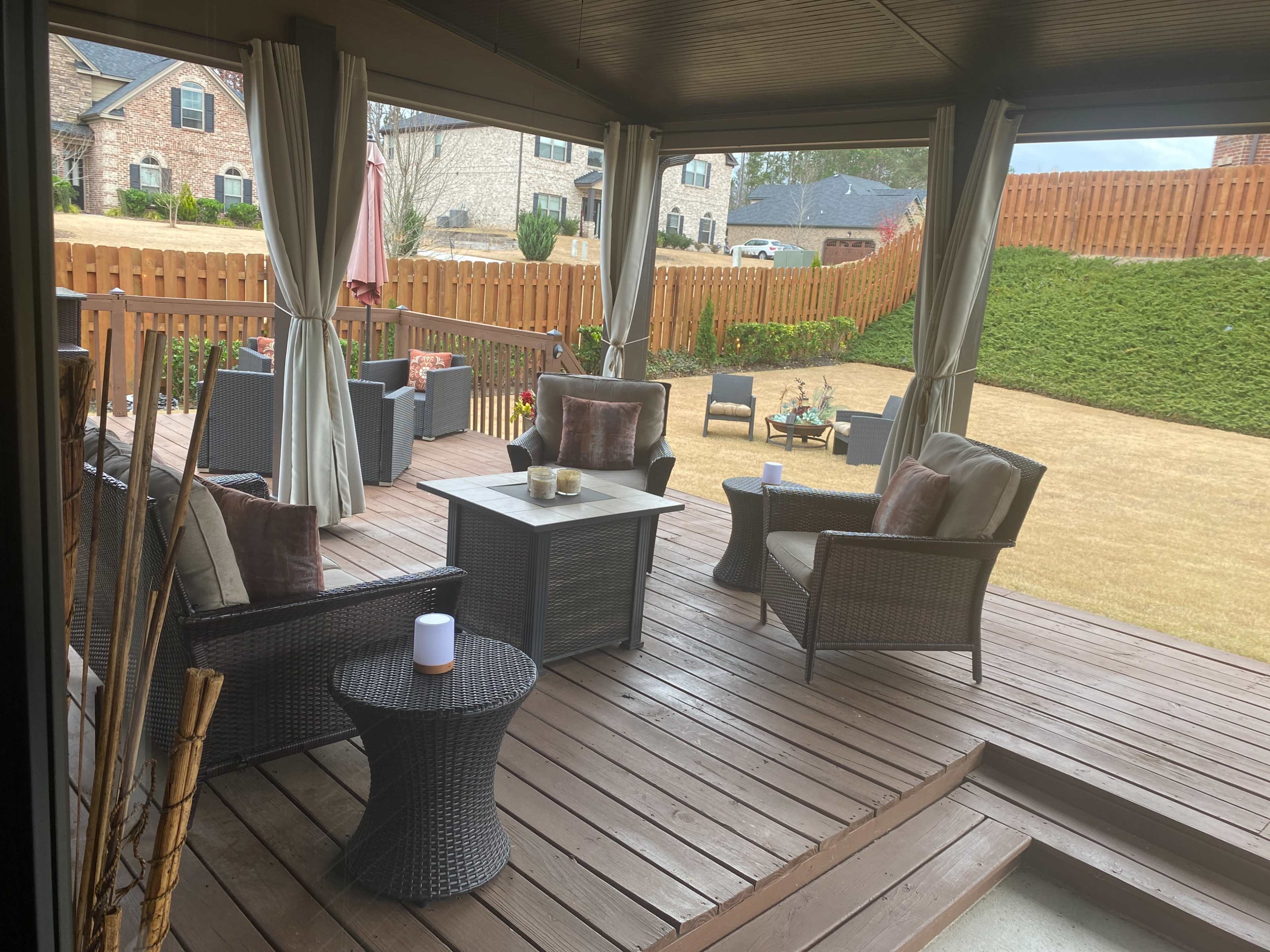 A covered outdoor seating area with wicker furniture, a wooden deck, and a fenced backyard.