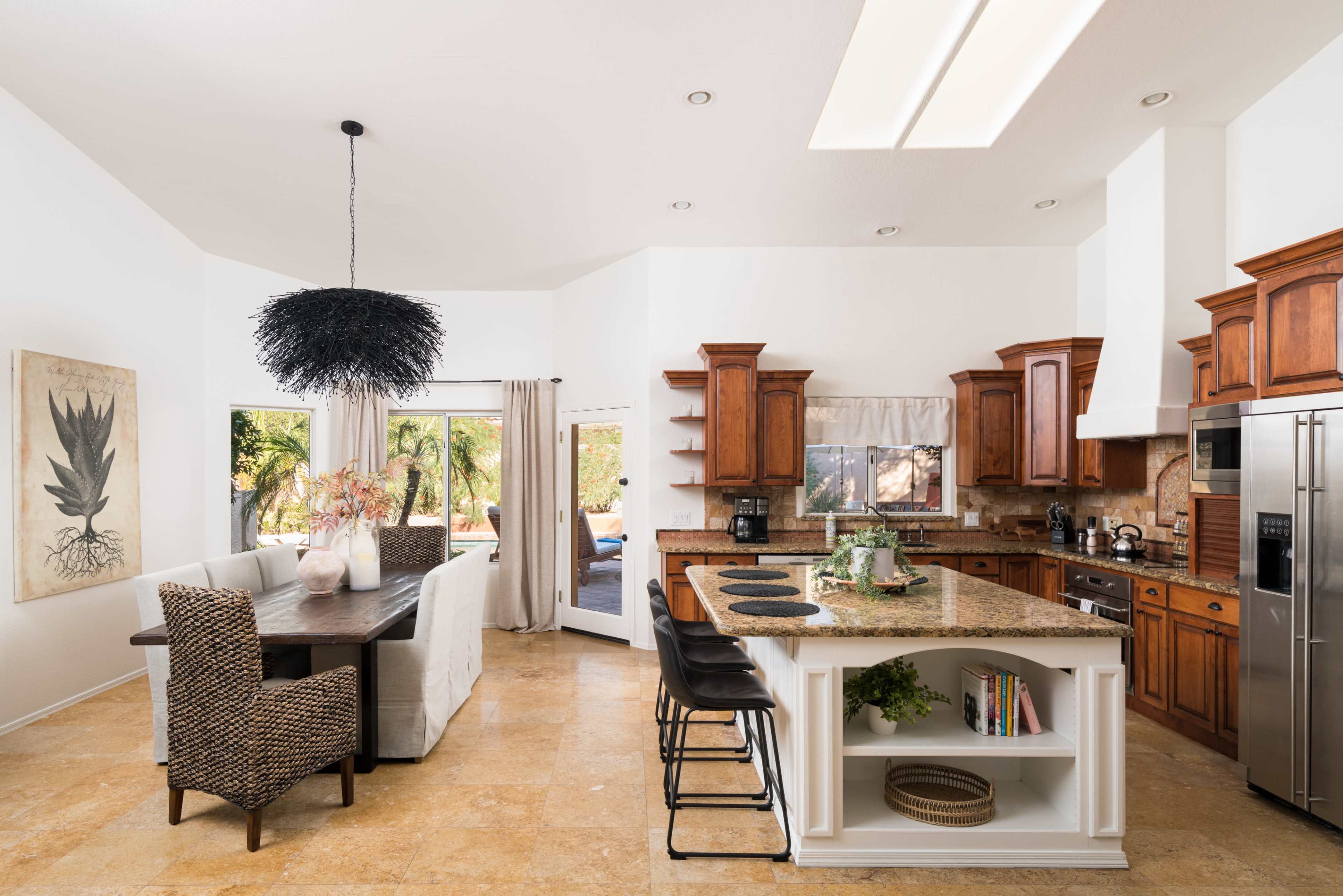 The image depicts a spacious kitchen with wooden cabinetry, a large island with bar stools, and a dining area featuring a textured pendant light and a table.