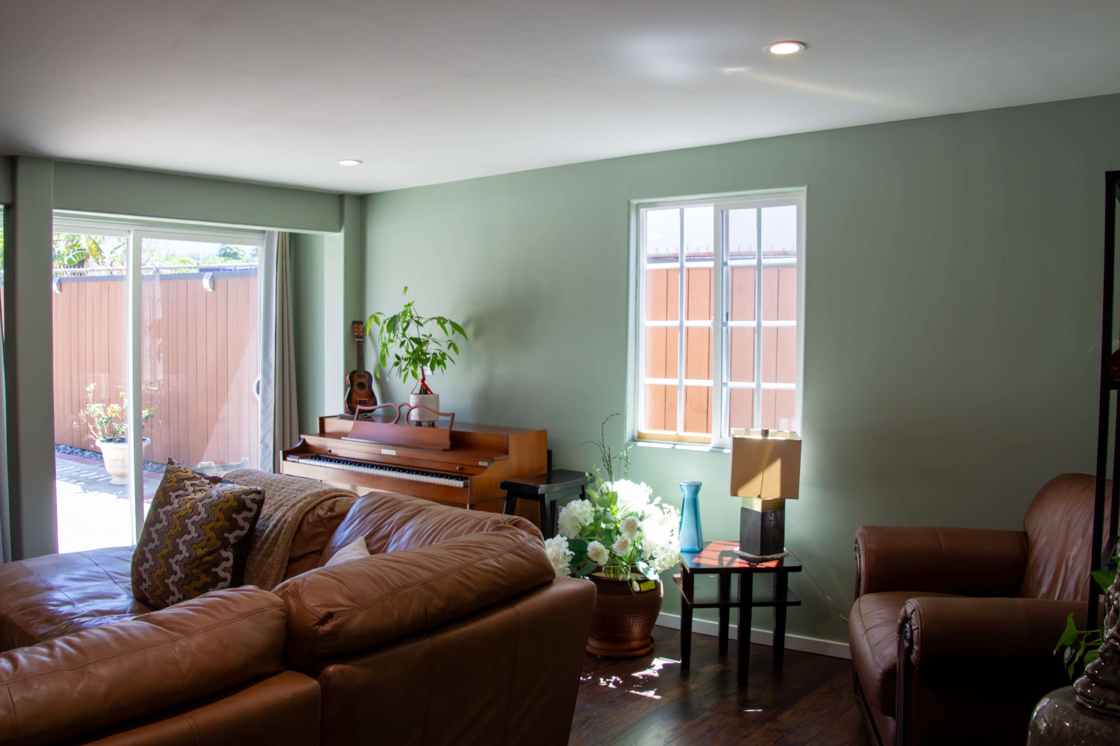 A living room features a brown leather sofa, an upright piano, and a large window with white frames.