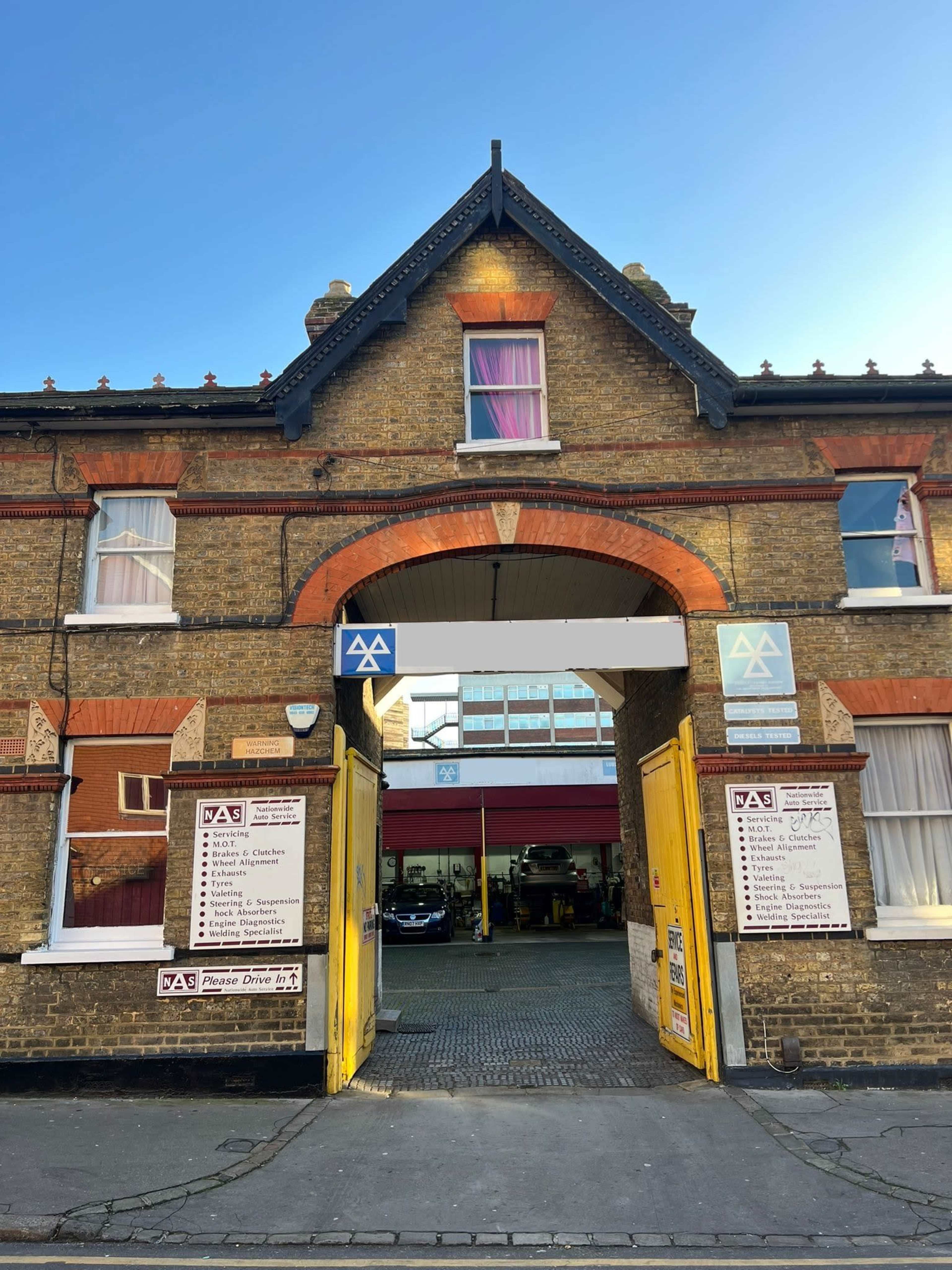 A brick building with a large arched entrance and yellow double doors, leading to a parking area, with signs on either side.