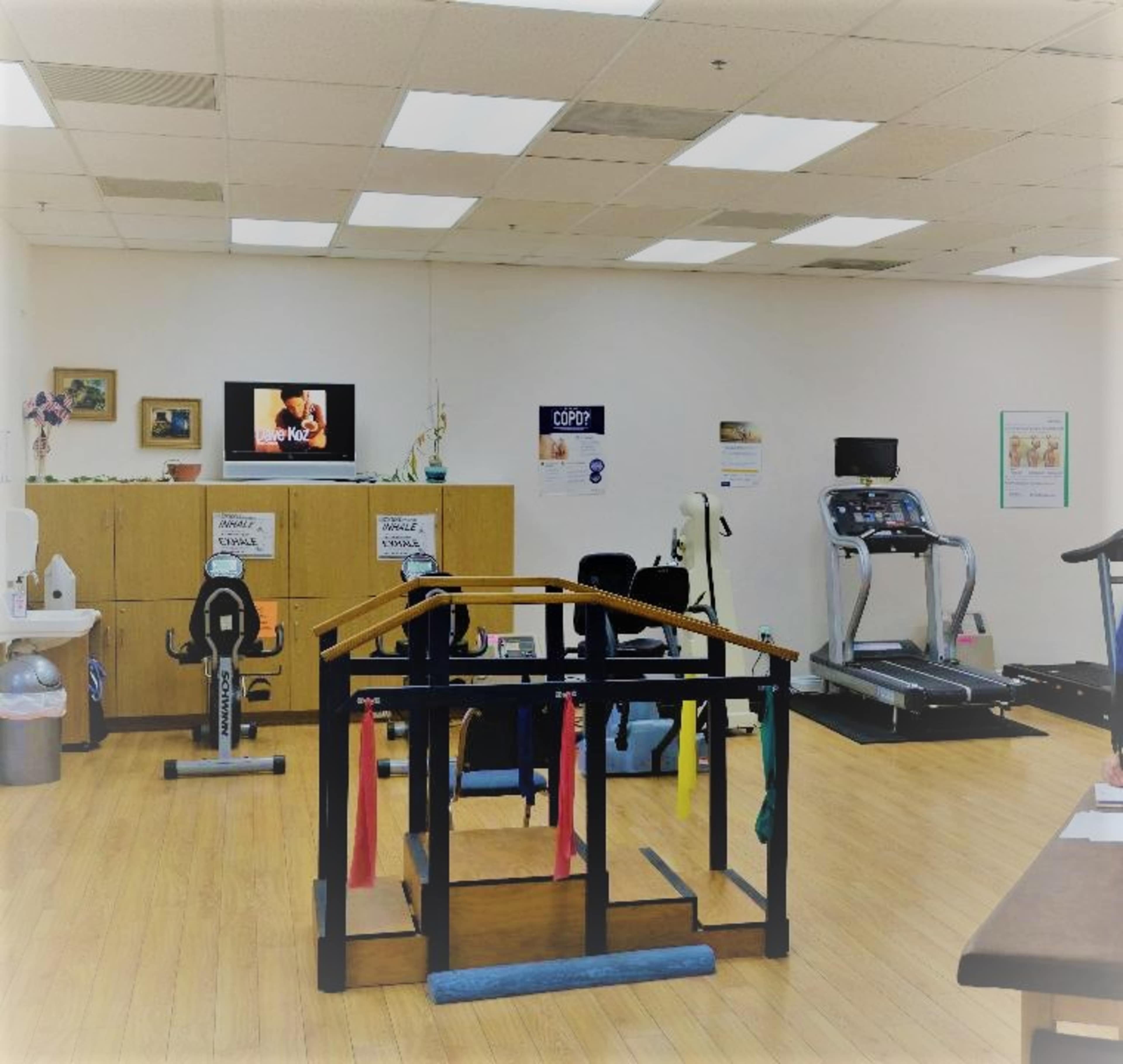 A fitness area with exercise equipment, including a treadmill, stationary bike, and a multi-use machine, situated in a well-lit room with wooden flooring.