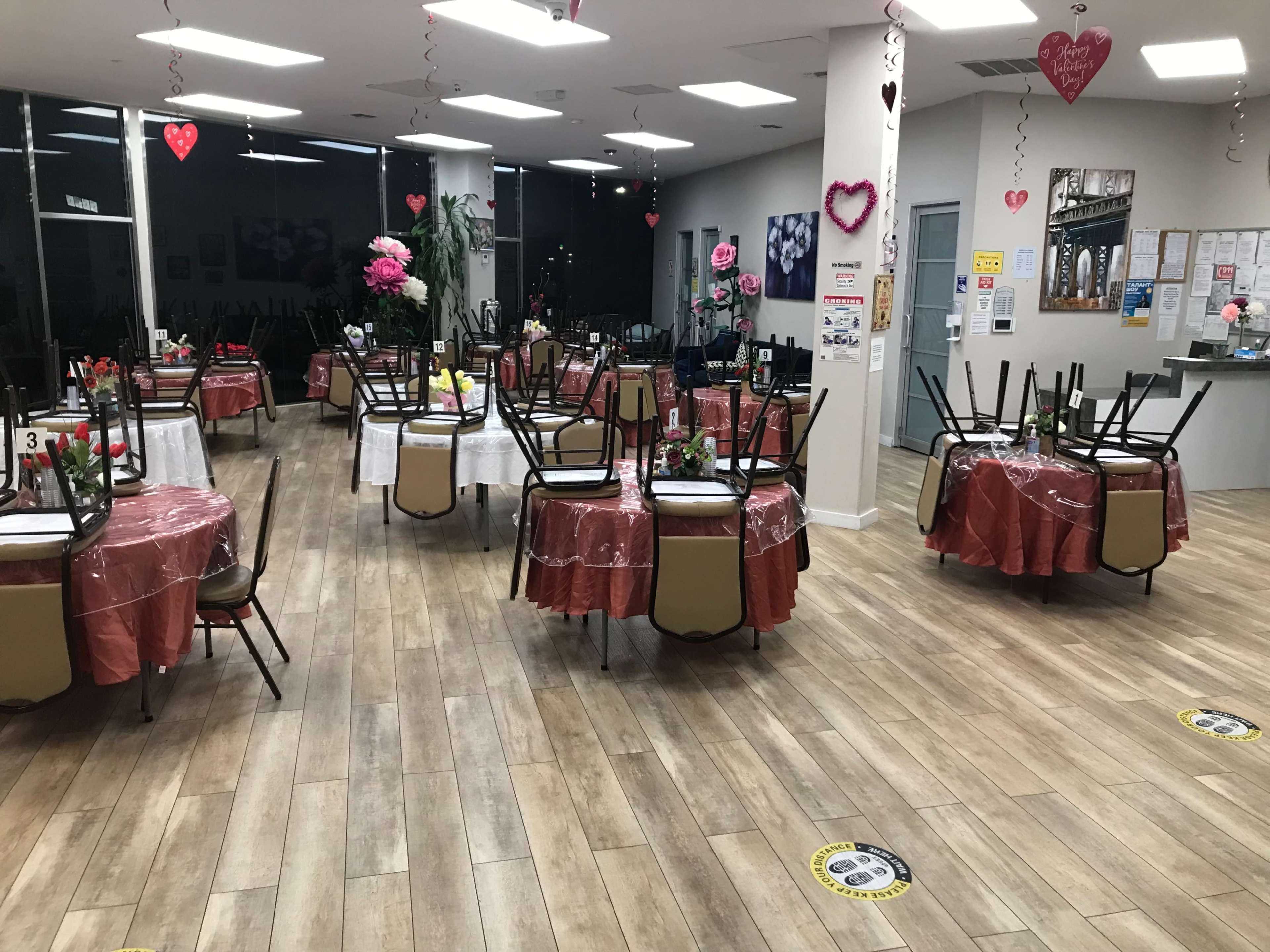 The restaurant features several round tables with chairs stacked on top, decorated with tablecloths and artificial flowers, set in a spacious area with a neutral color scheme.