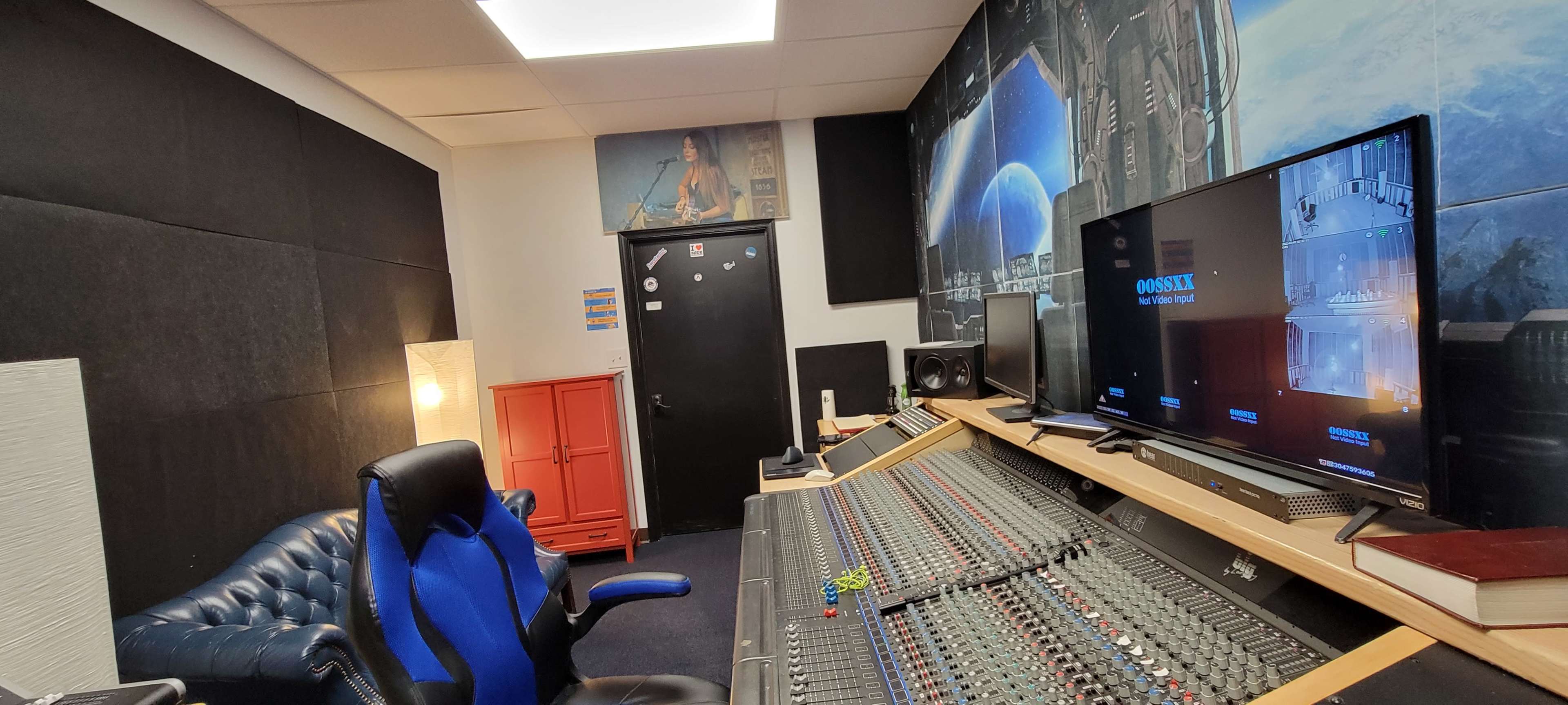 A recording studio with a large mixing console, a computer monitor displaying audio software, and a blue leather chair in a room featuring soundproofing panels on the walls.
