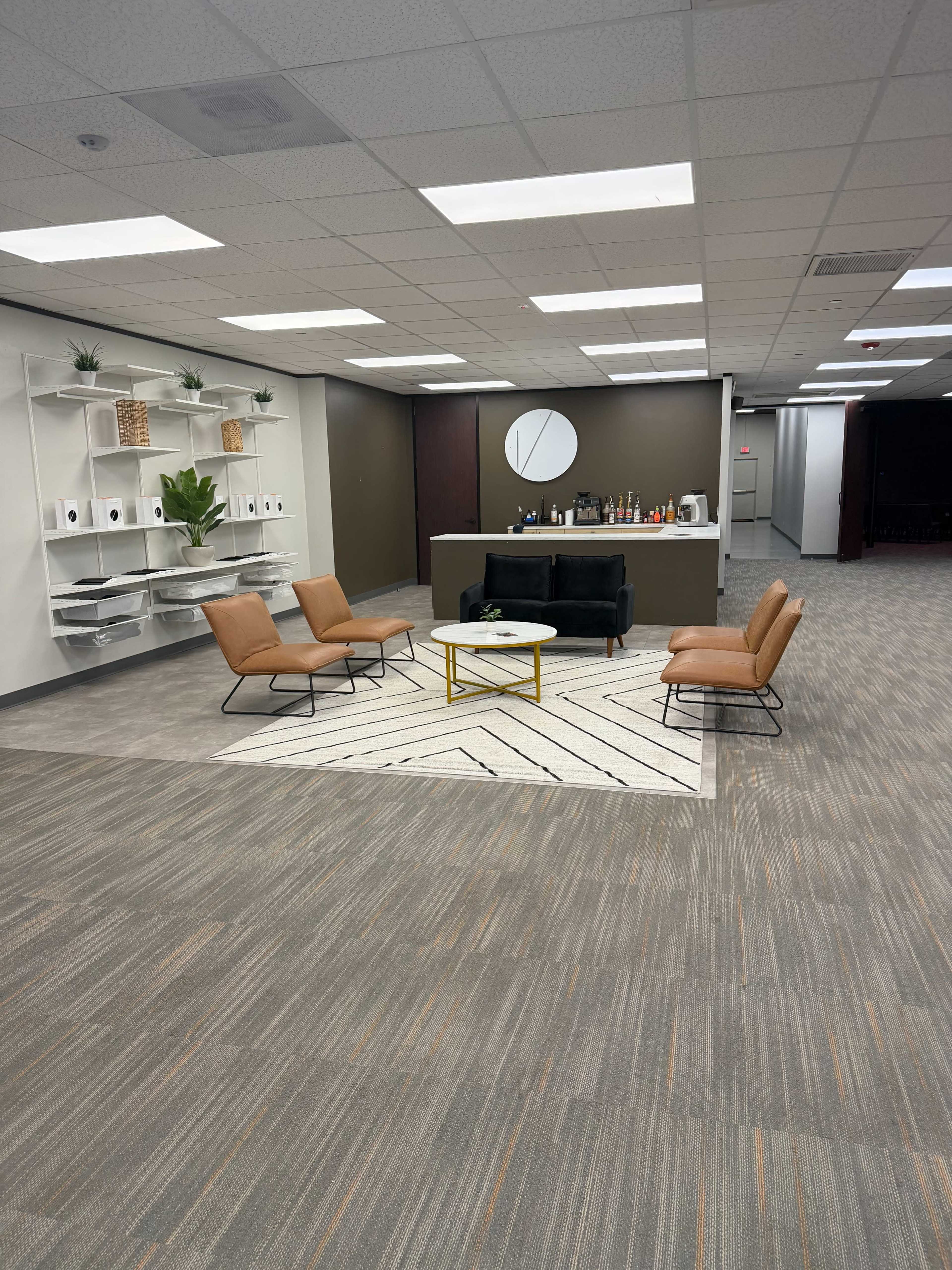 The image shows a modern office lounge area with four chairs surrounding a small table on a patterned rug, a bar area in the background, and shelves with decorative items on the wall.