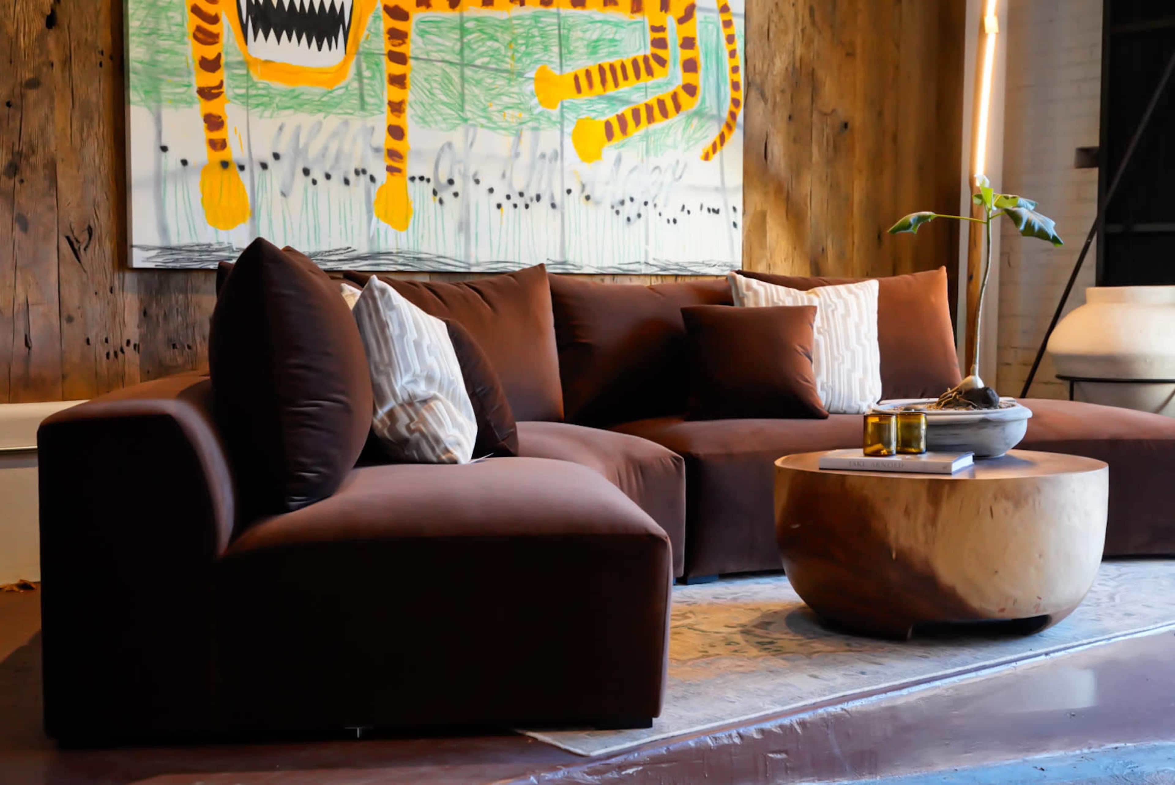 The image shows a modern living room with a brown sectional sofa, a round wooden coffee table, and a piece of abstract wall art featuring a colorful tiger.