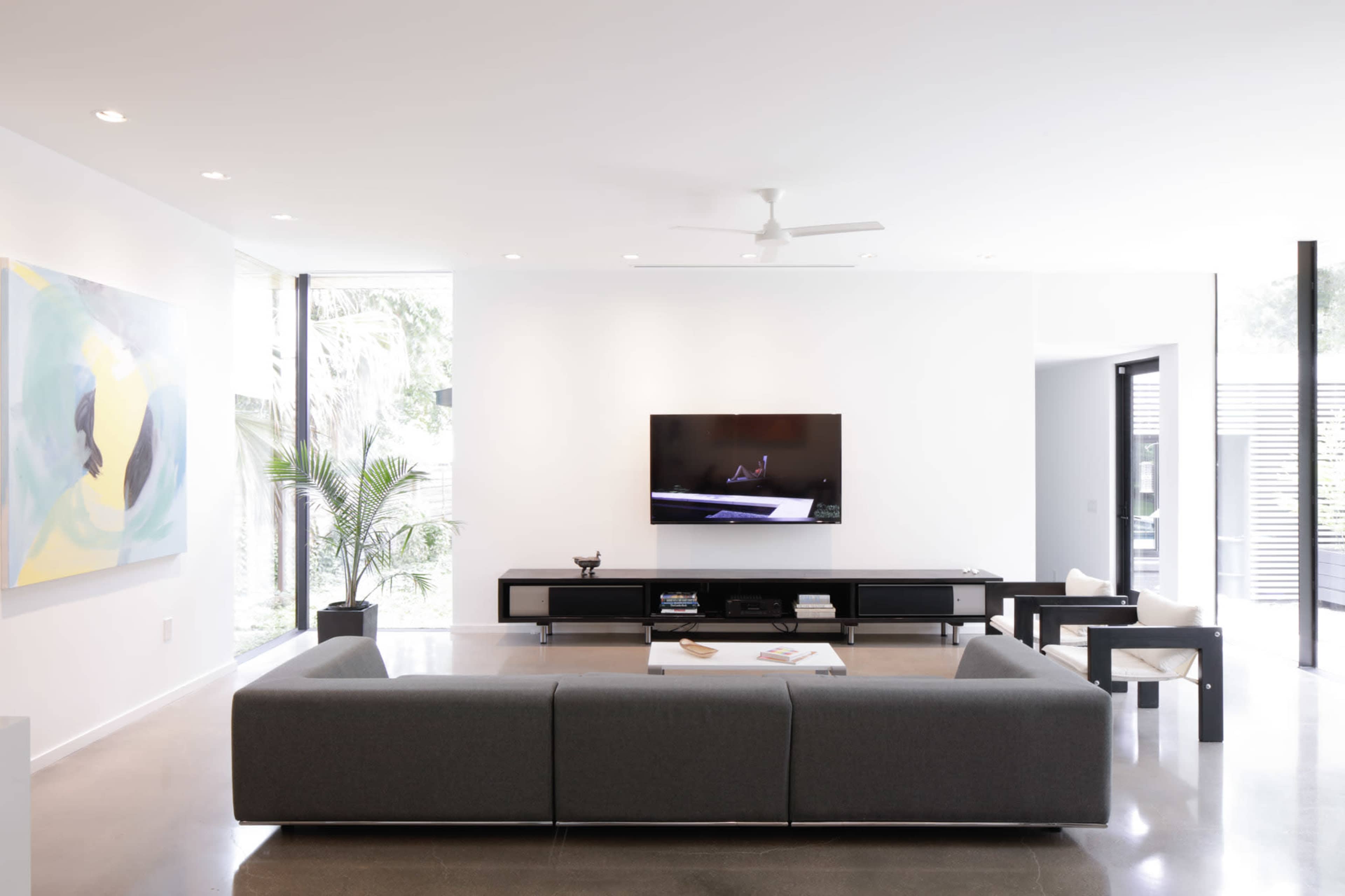 A modern living room features a gray sofa facing a wall-mounted television, with a black media console and large windows allowing natural light to enter.