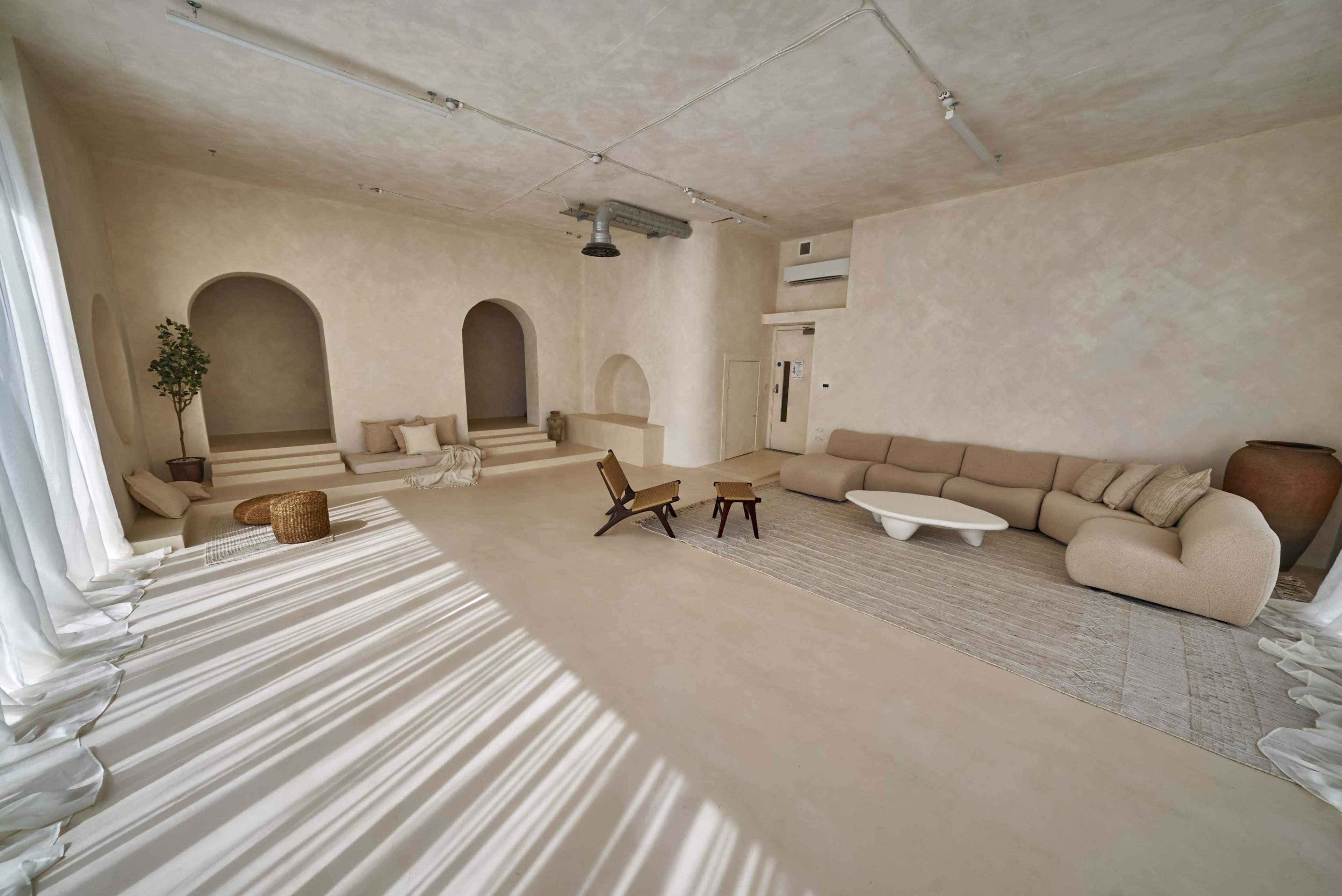 The image shows a spacious, minimalist interior with soft, neutral tones, featuring a curved sofa, a coffee table, and decorative elements such as a potted plant and woven items.