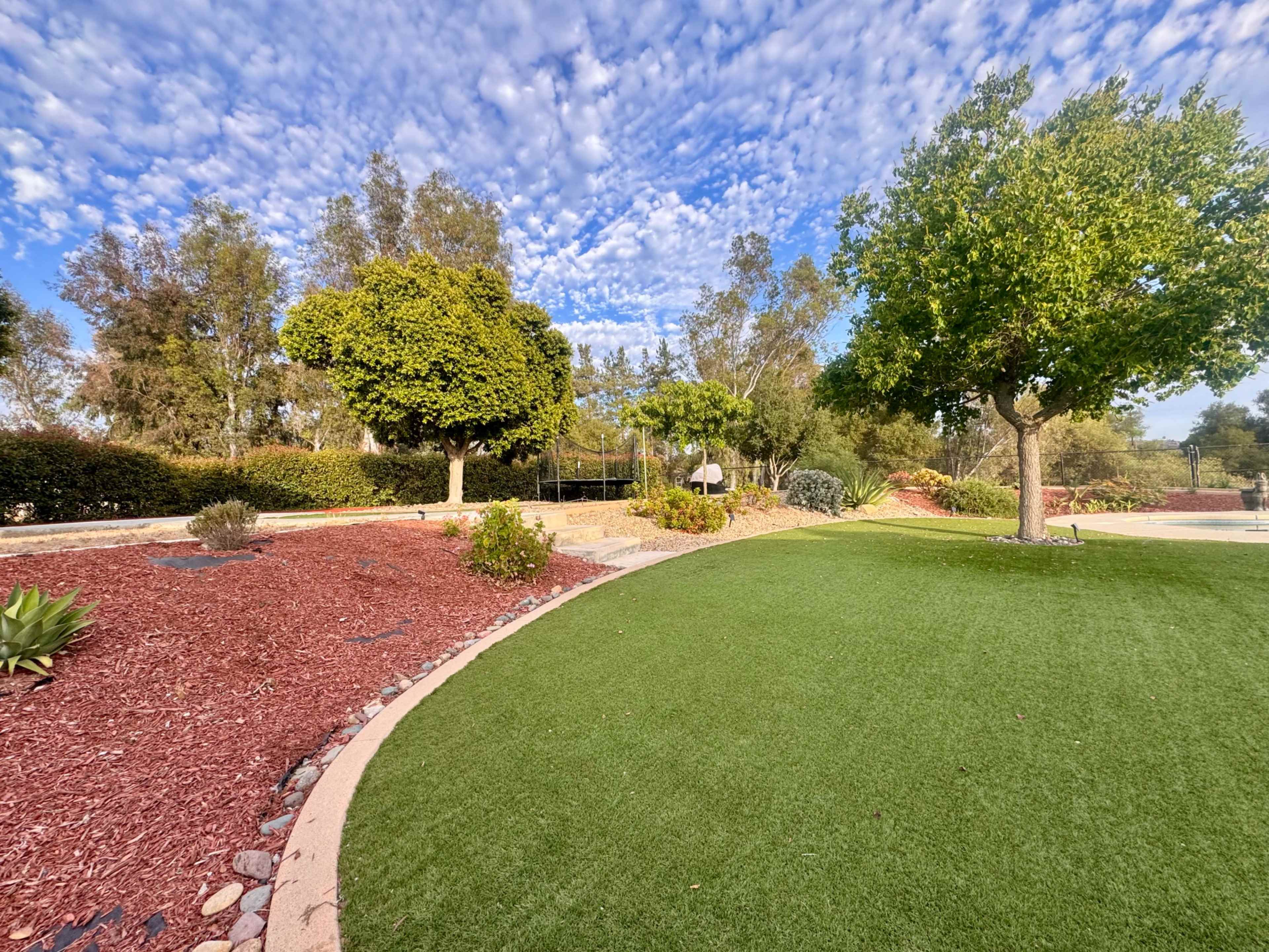 The image depicts a landscaped park area featuring green grass, gravel pathways, and trees under a cloudy blue sky.