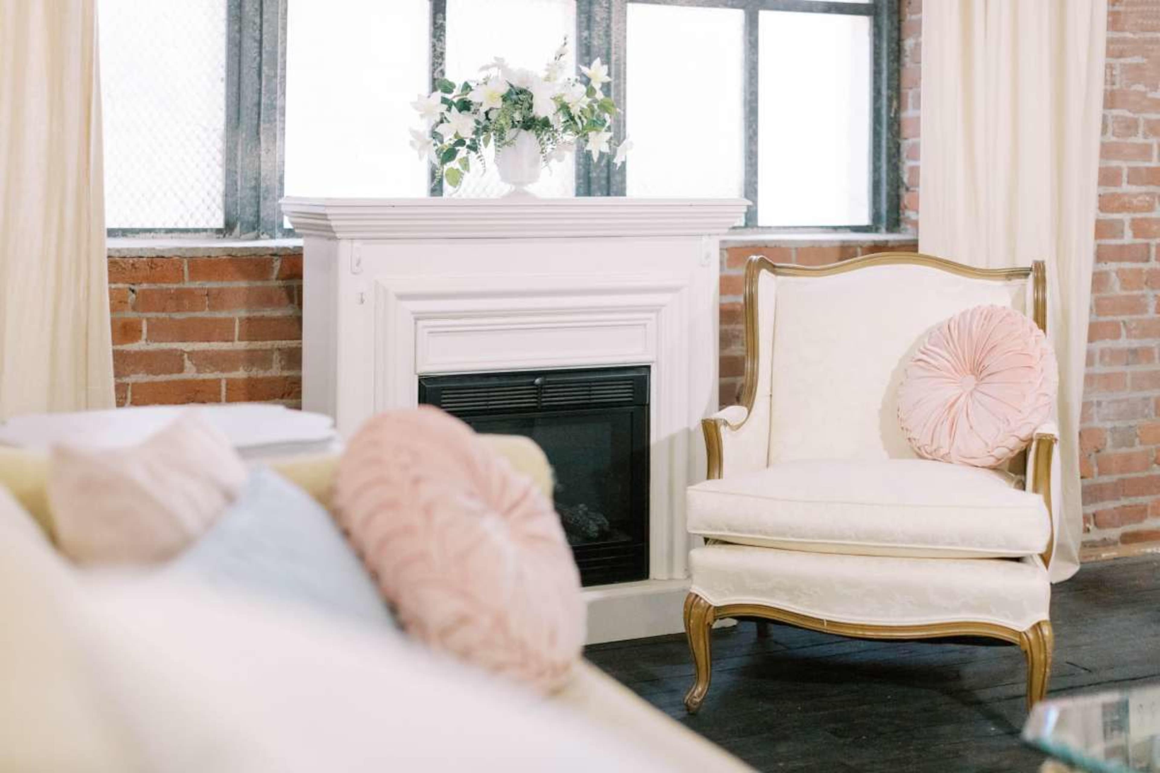 The image shows a cozy living room with a white chair featuring a pink decorative pillow next to a white fireplace, all set against a backdrop of exposed brick walls and large windows.