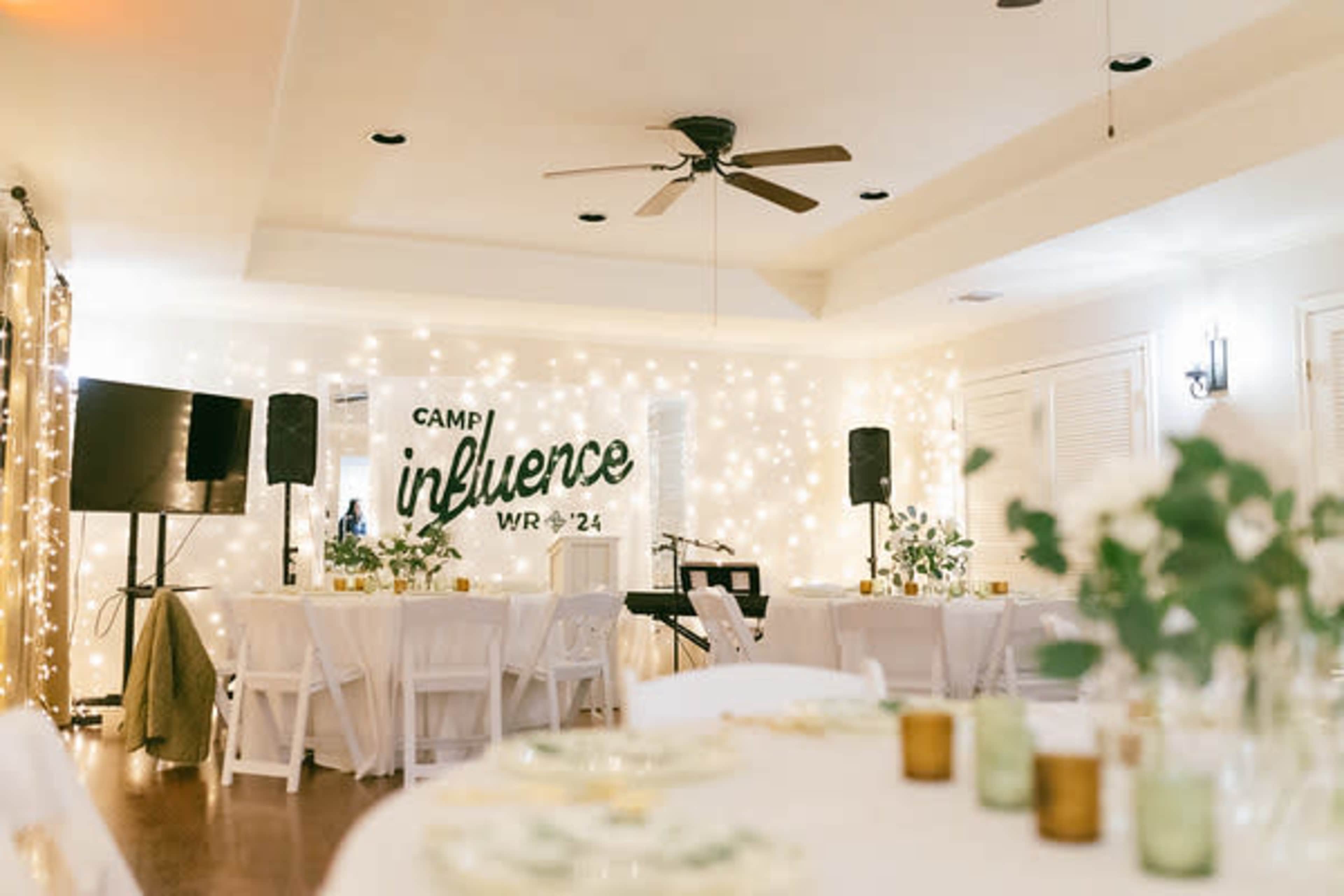 A decorated indoor event space featuring tables, string lights, and a backdrop with the words "Camp Influence WR 24."