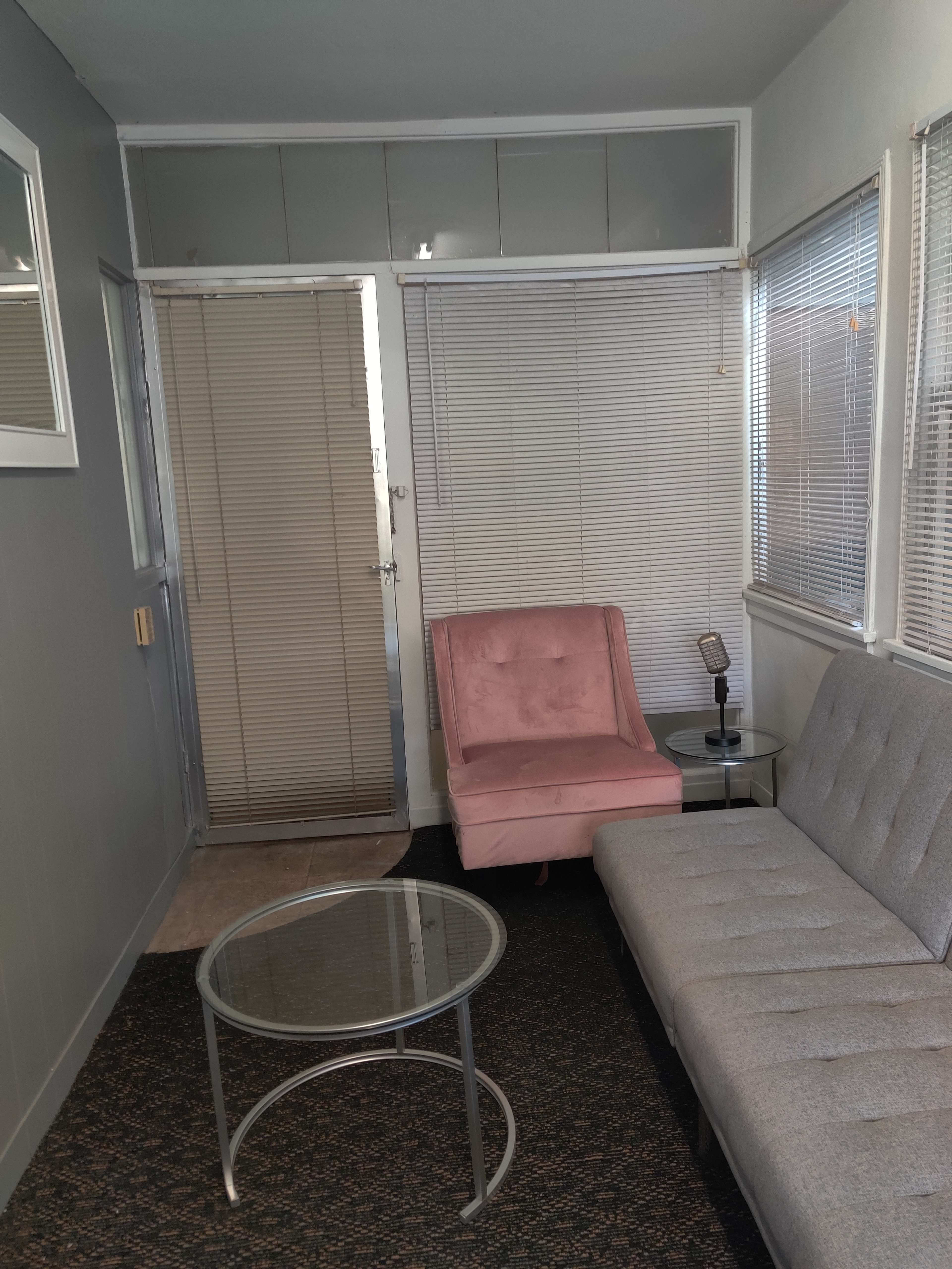 A small living room with a grey couch, a pink armchair, a round glass coffee table, and a door leading outside.