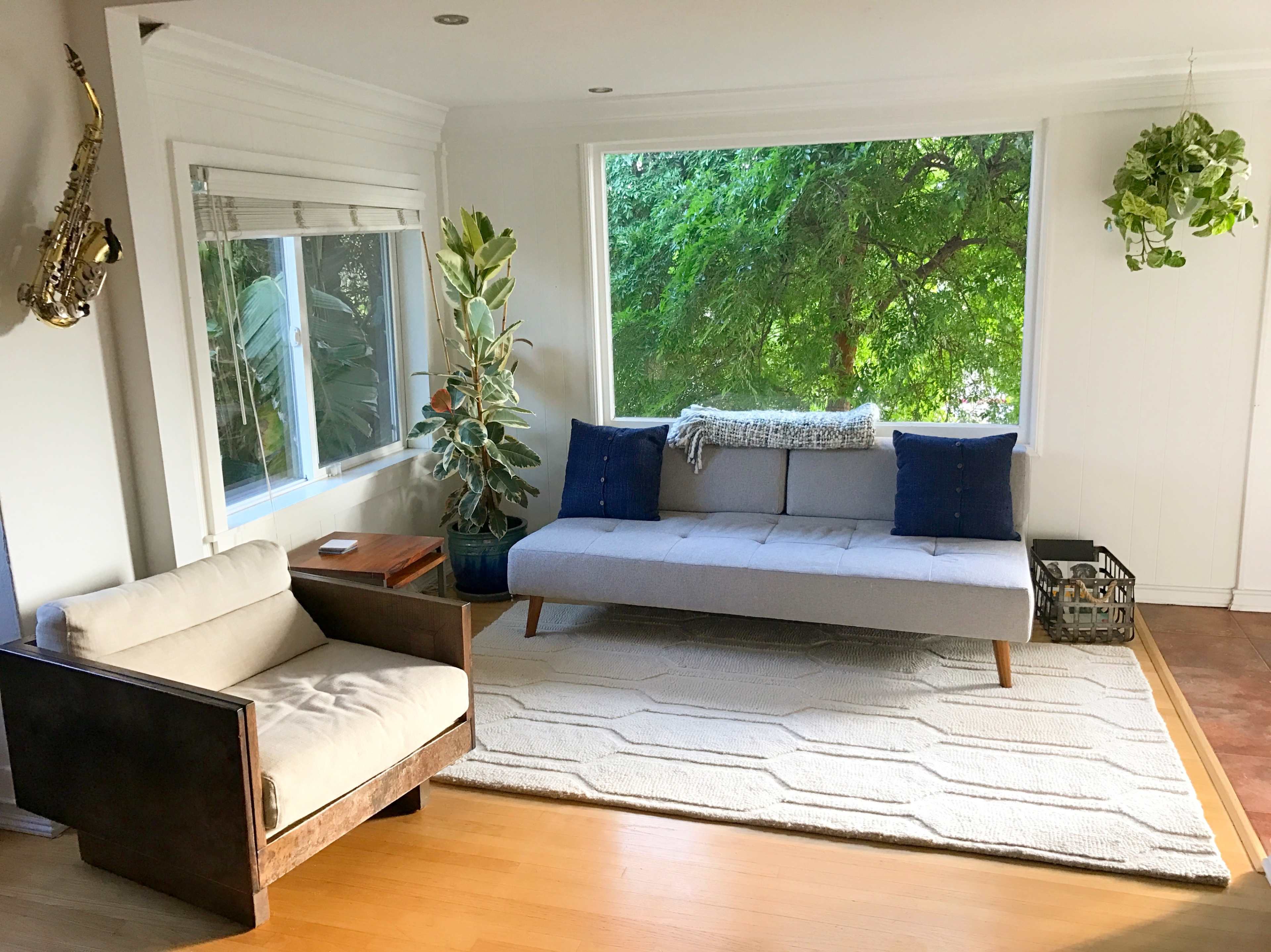 A modern living room features a gray sofa, a wooden chair, a large window with greenery outside, and a light rug on the floor.
