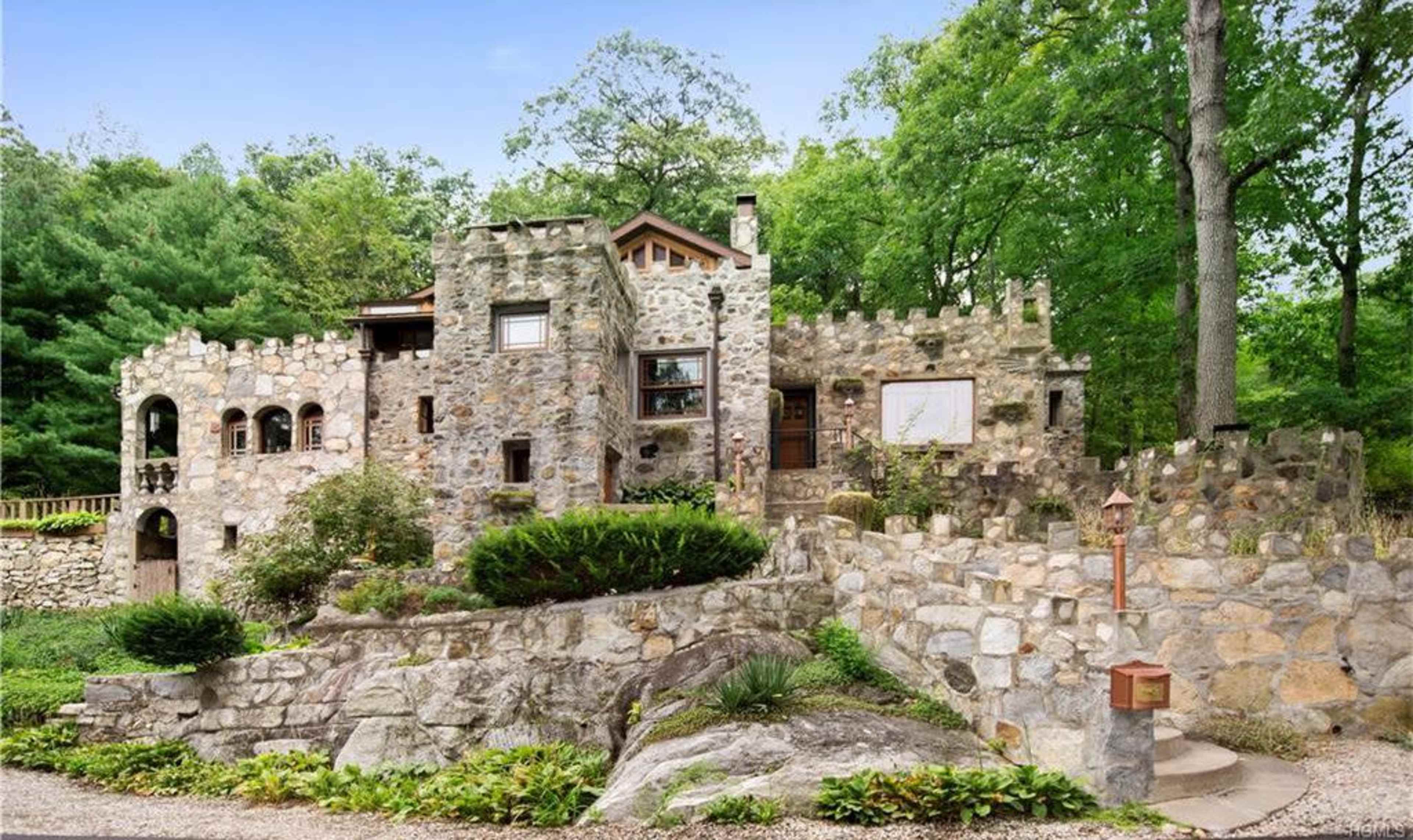 A stone house designed with castle-like features, surrounded by lush greenery and a landscaped pathway.