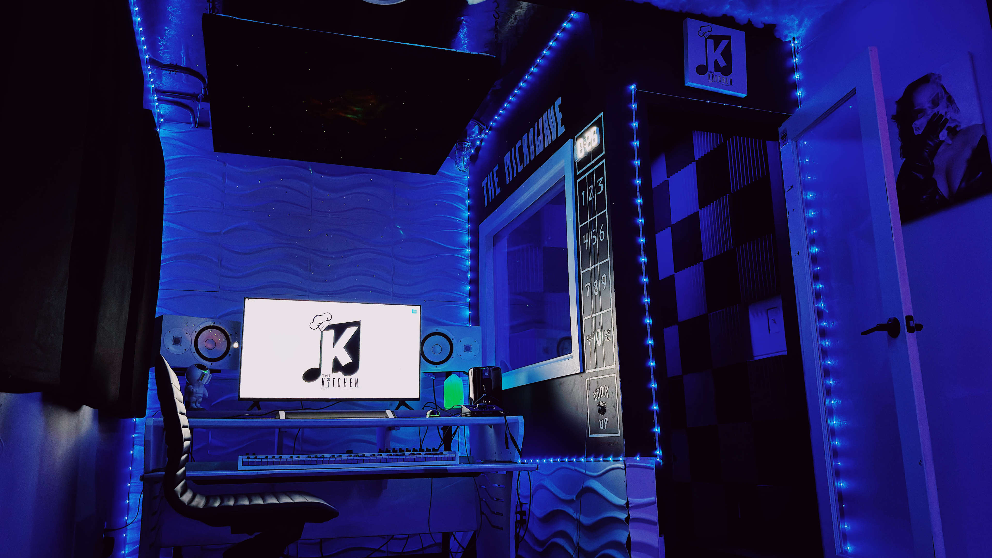 The image shows a music studio with blue LED lighting, a computer desk, monitors, and soundproofing panels on the walls.