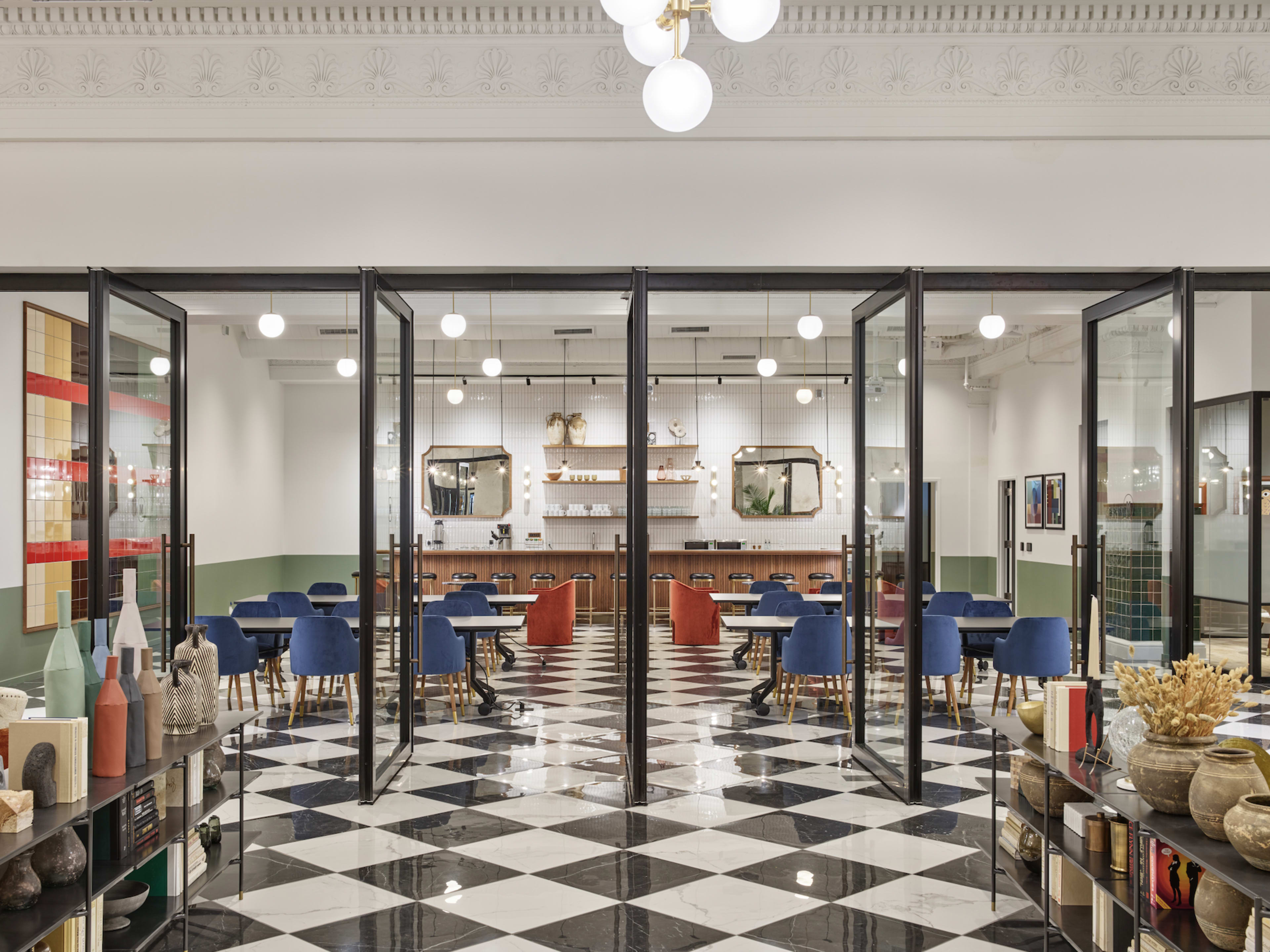 A modern interior space with a checkered black and white floor, featuring blue chairs and wooden tables, separated by glass doors from another area that includes decorative vases and artwork.