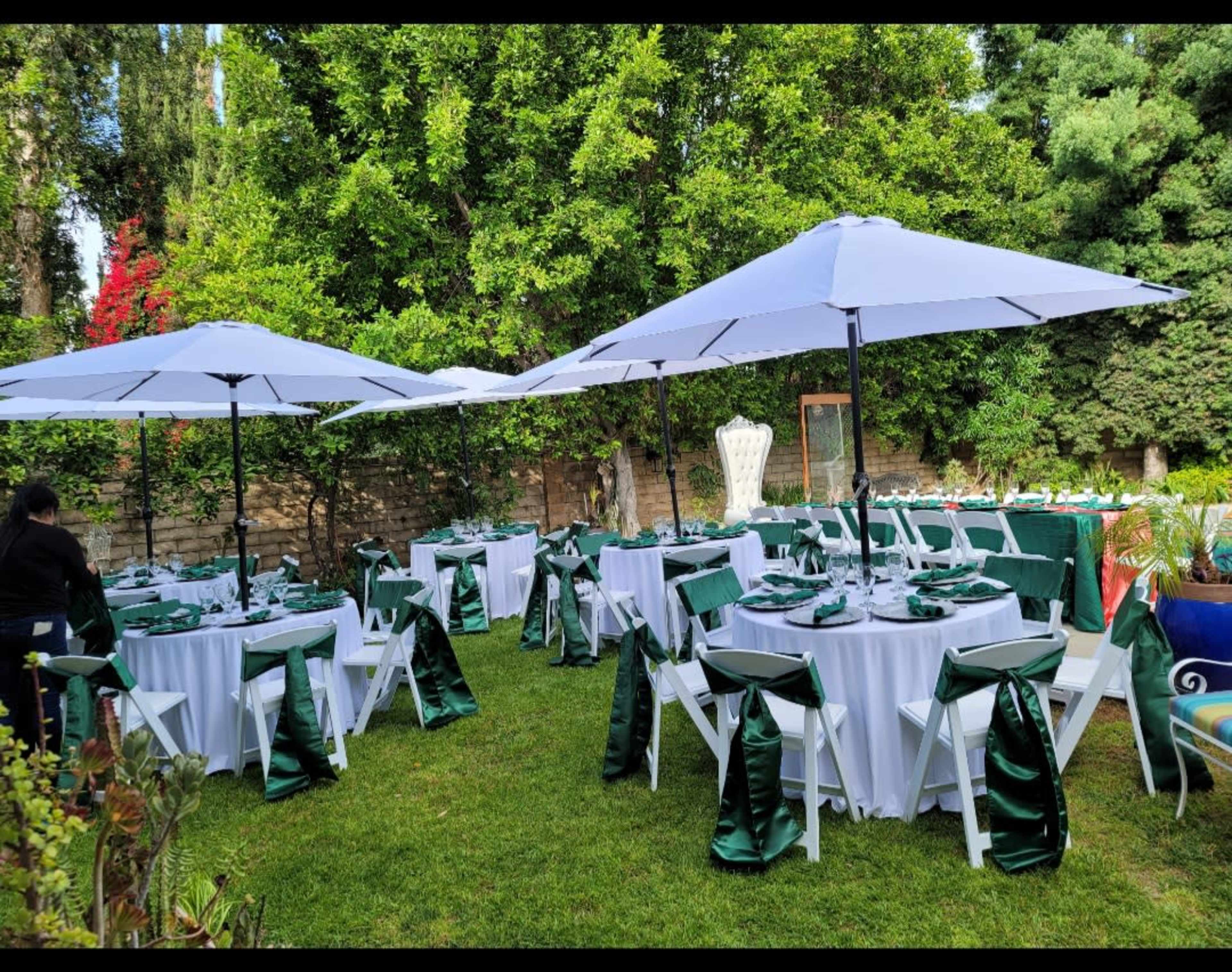 A neatly arranged outdoor event space features tables covered with white linens and decorated with green accents, shaded by large white umbrellas.