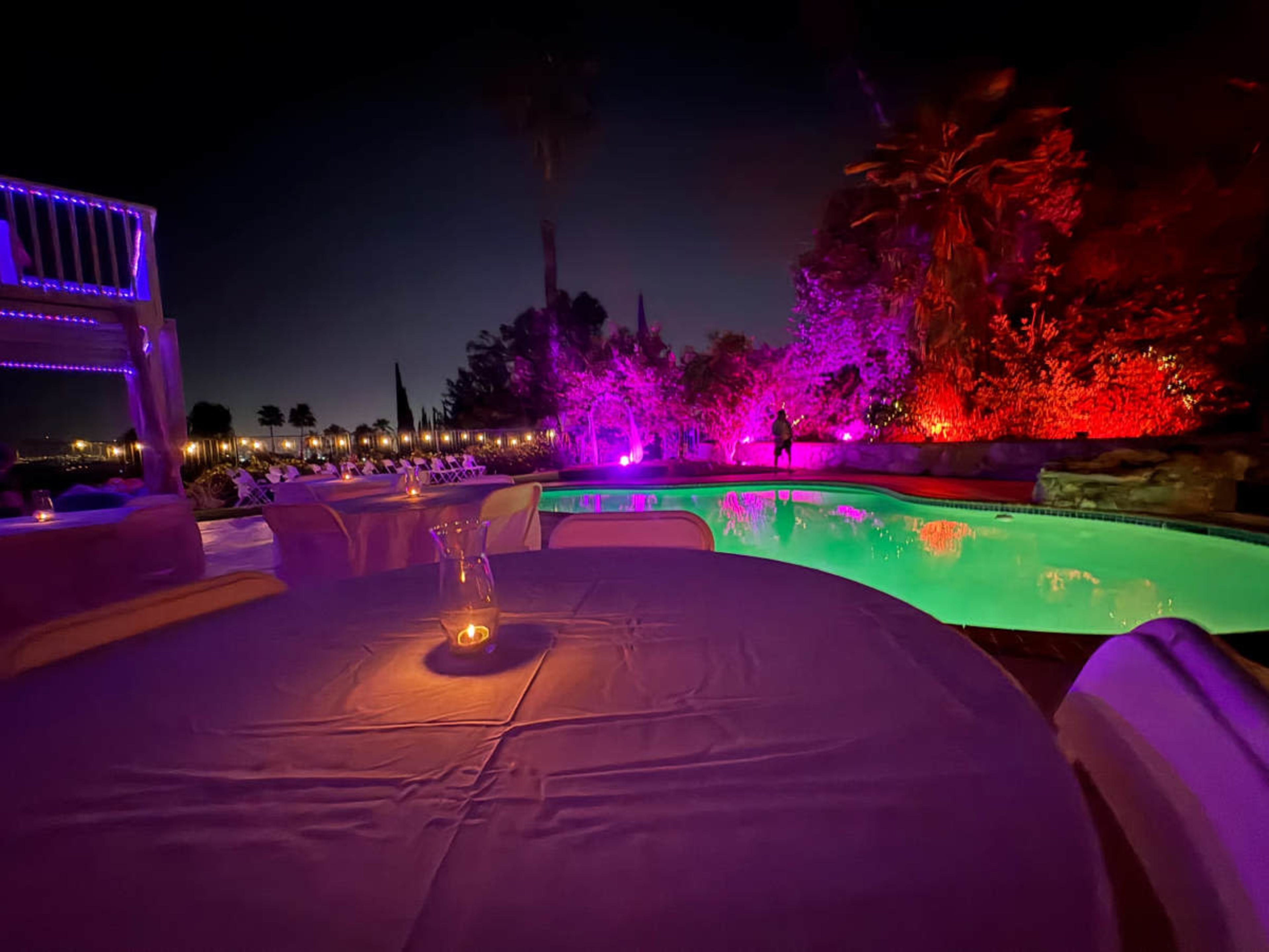 A table is set by a illuminated swimming pool surrounded by colorful lighting and palm trees at night.