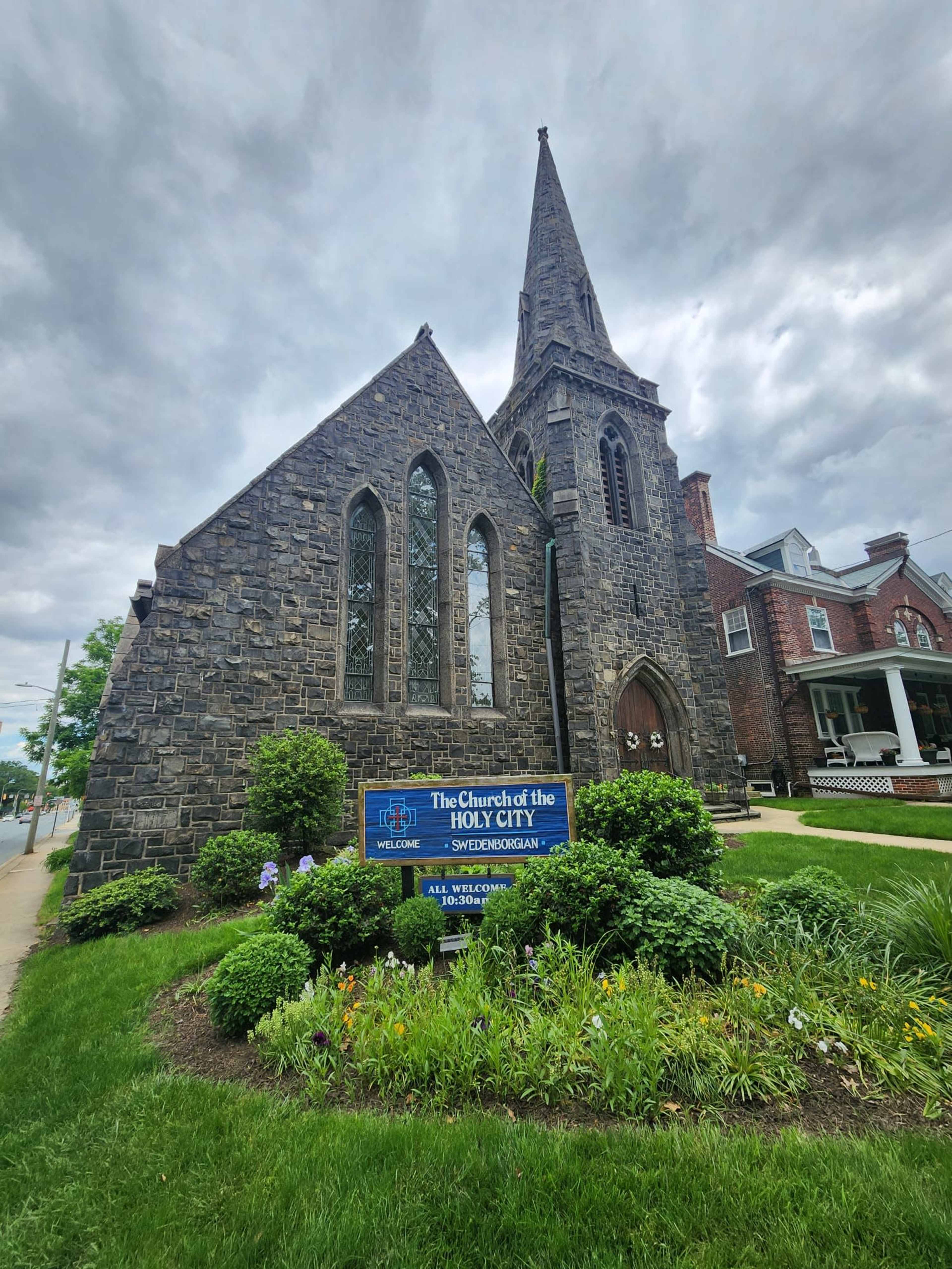 The image shows a stone church with a tall steeple, surrounded by greenery and a sign indicating it is the Church of the Holy City.