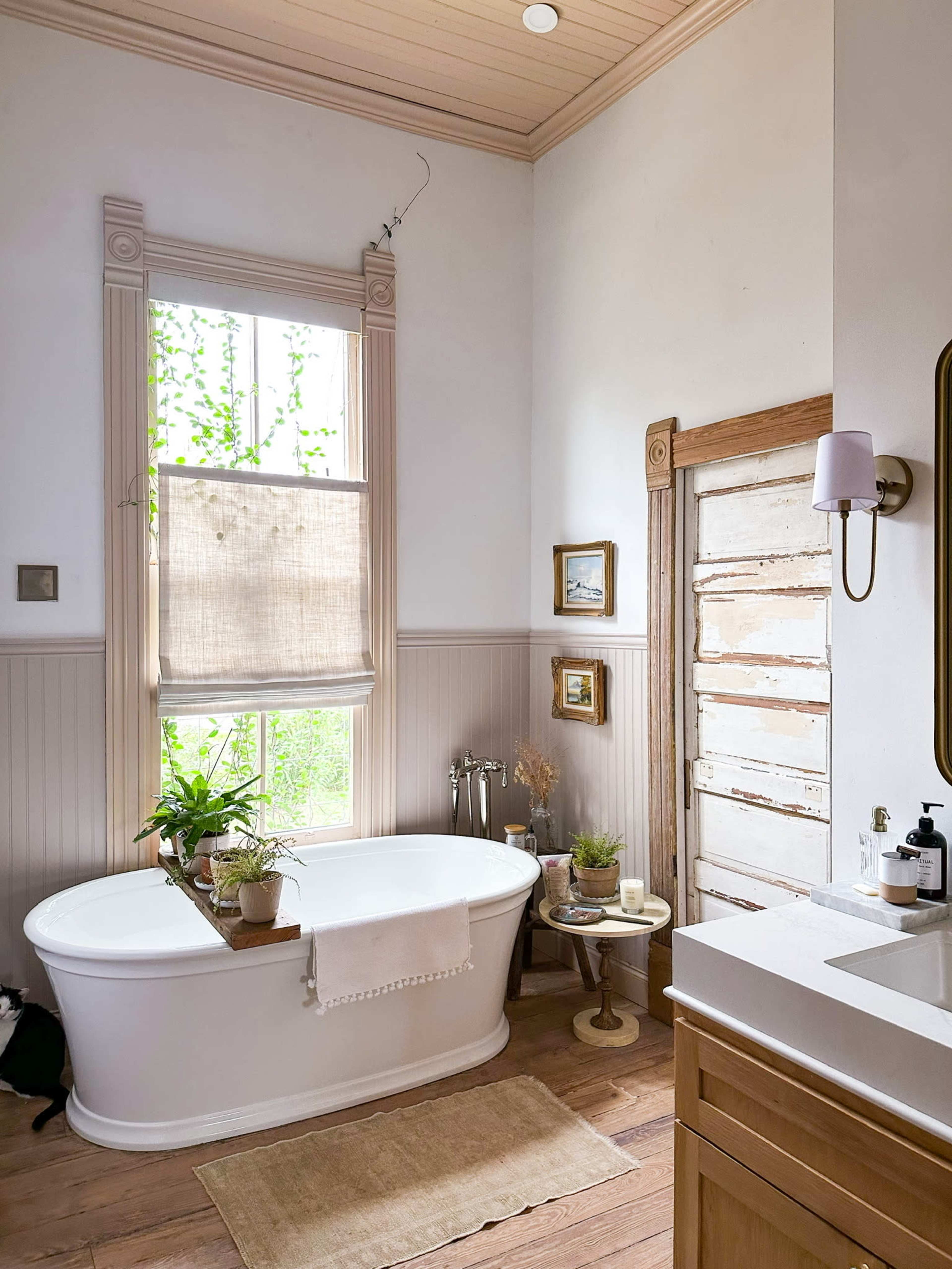 A bright bathroom featuring a freestanding bathtub, a wooden door, and a window covered with a woven shade, surrounded by plants and decorative items.