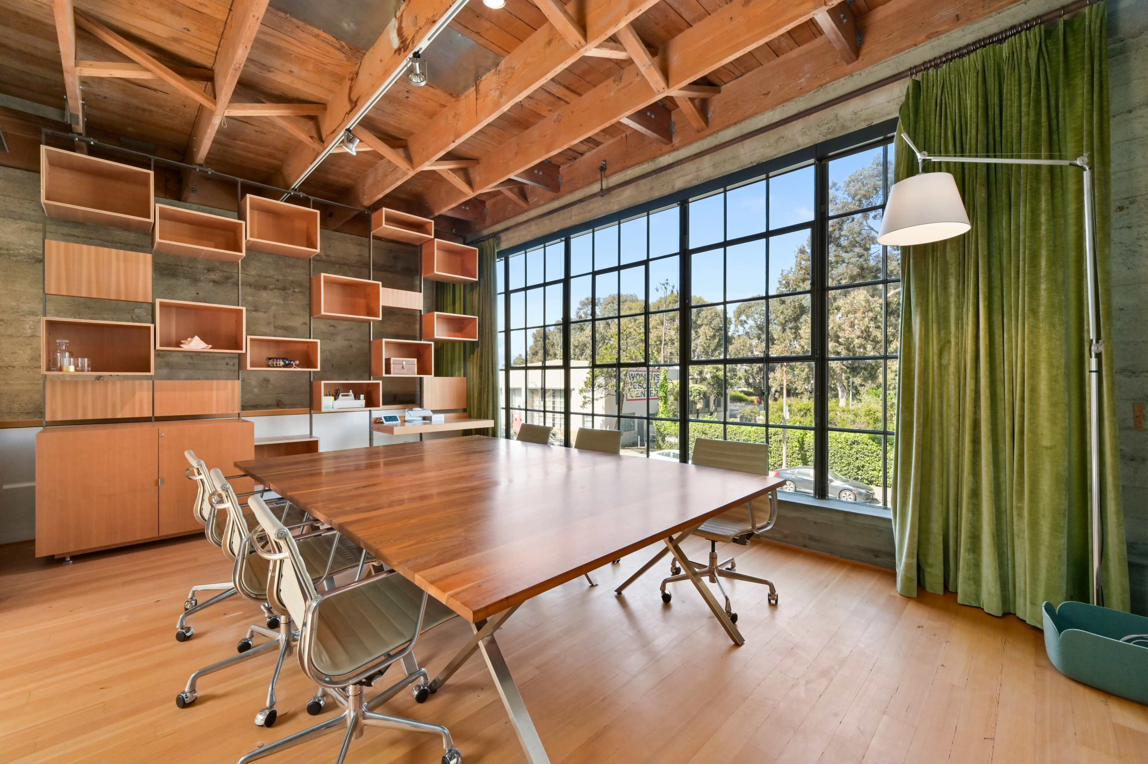 A modern conference room with a large wooden table, six metal chairs, and a large window flanked by green curtains, surrounded by shelving and a minimalist decor.