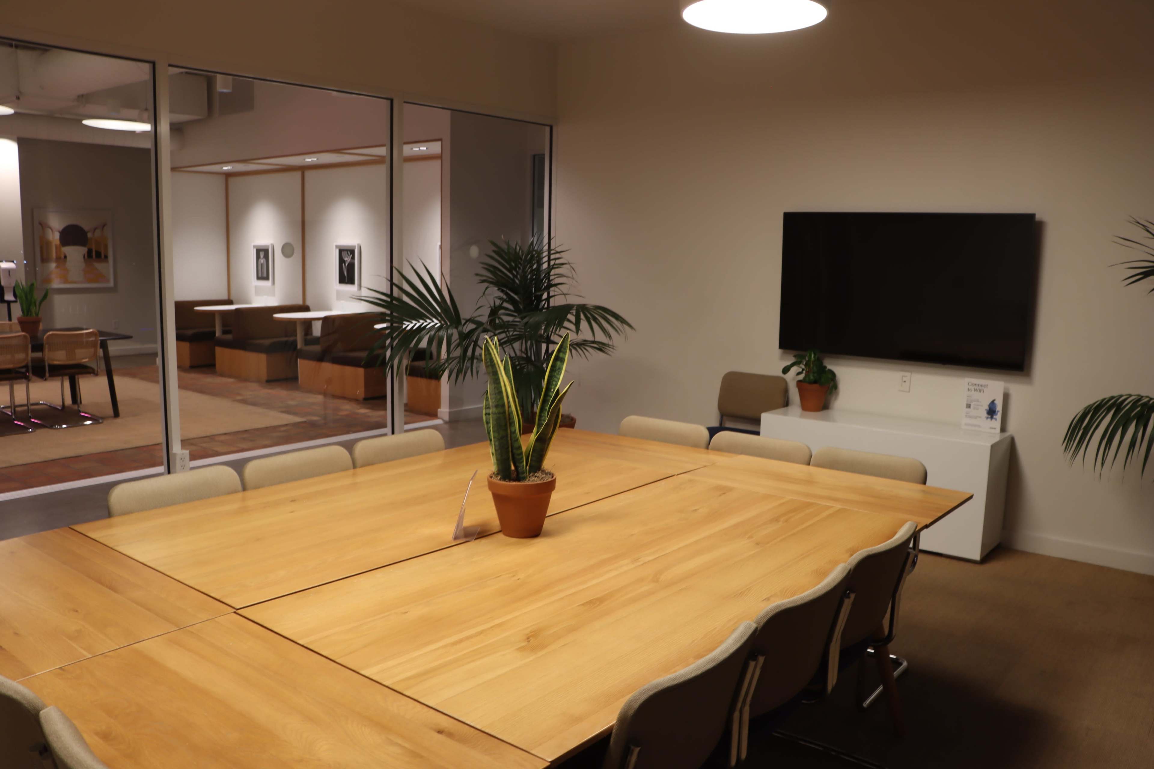 A rectangular wooden conference table is surrounded by beige chairs, with a potted plant in the center and a television mounted on the wall in a well-lit meeting room.