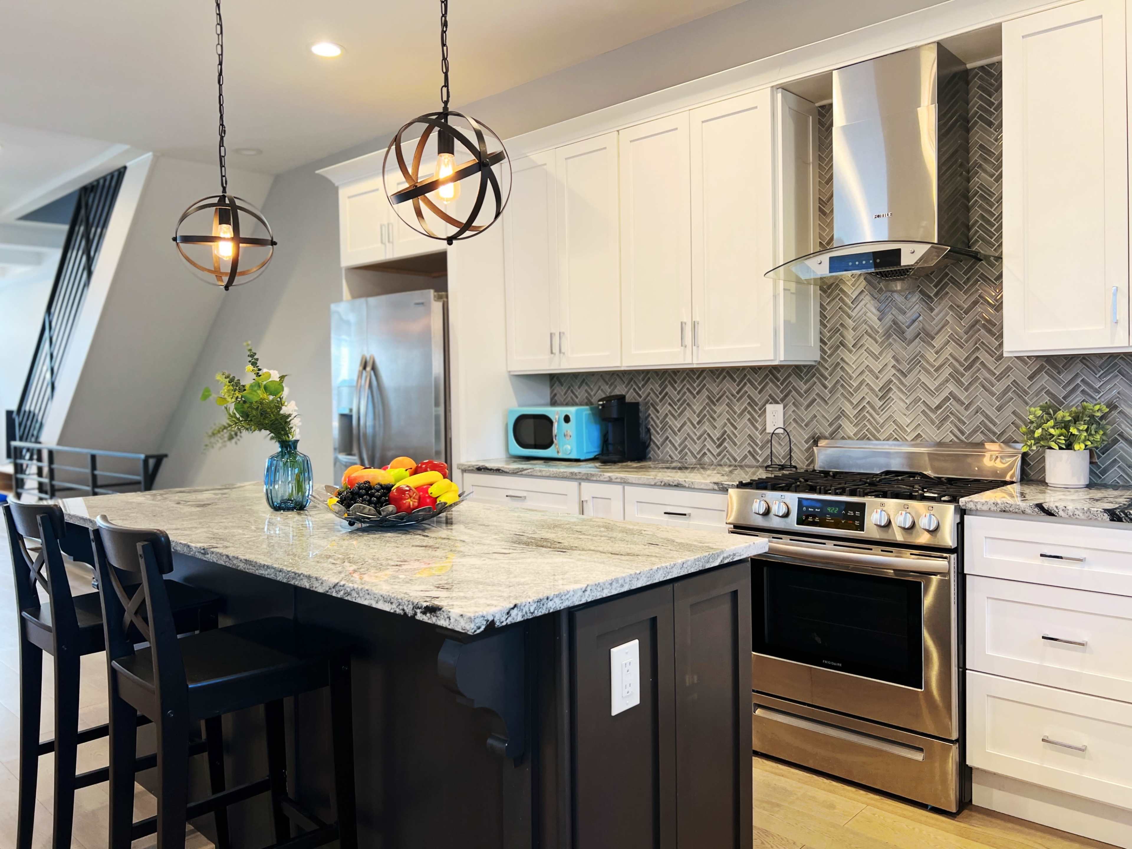 A modern kitchen with white cabinetry, stainless steel appliances, a gray and white herringbone backsplash, and a central island with bar stools and a fruit bowl.