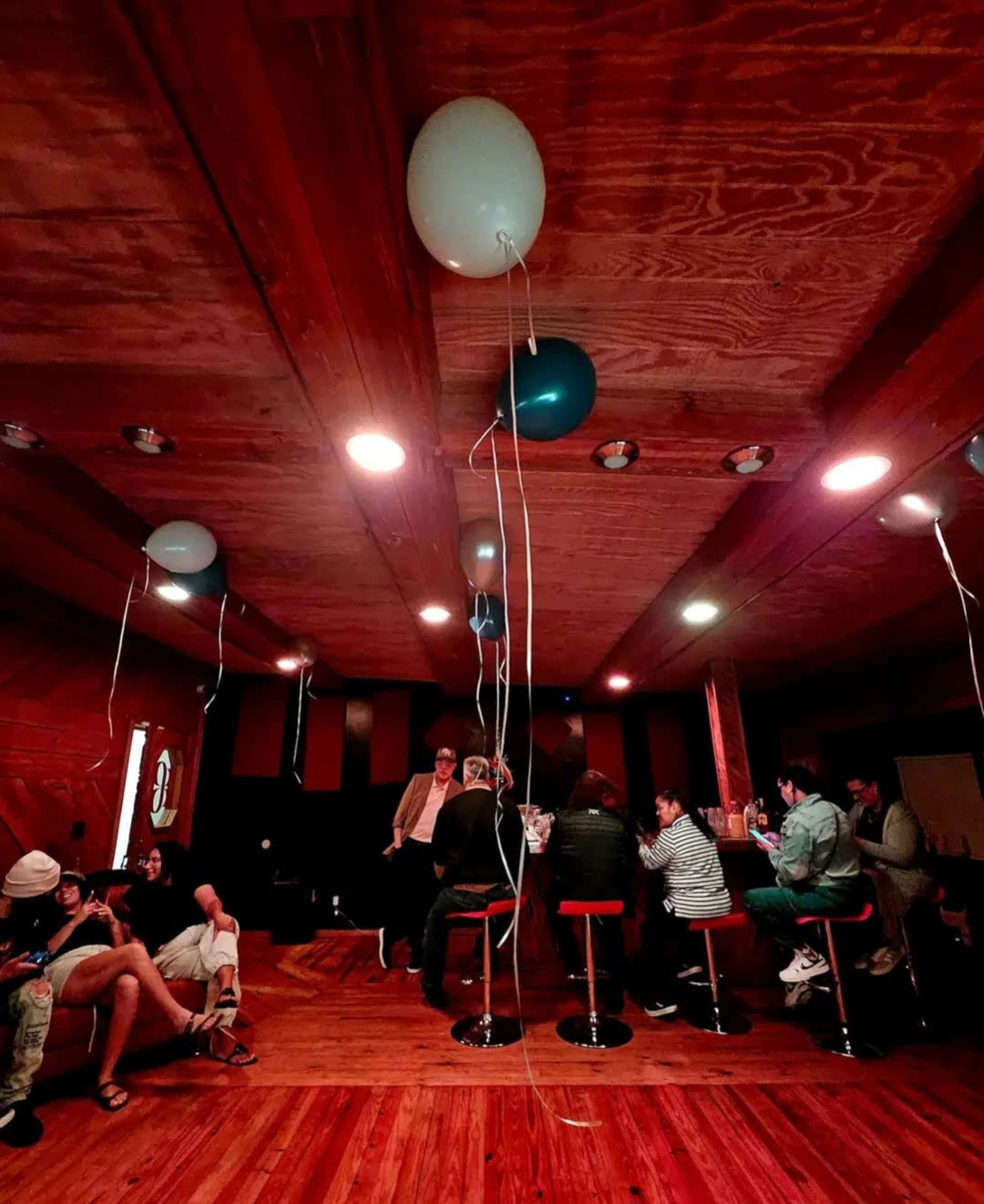 A wooden interior room with people seated at tables, decorated with balloons hanging from the ceiling.