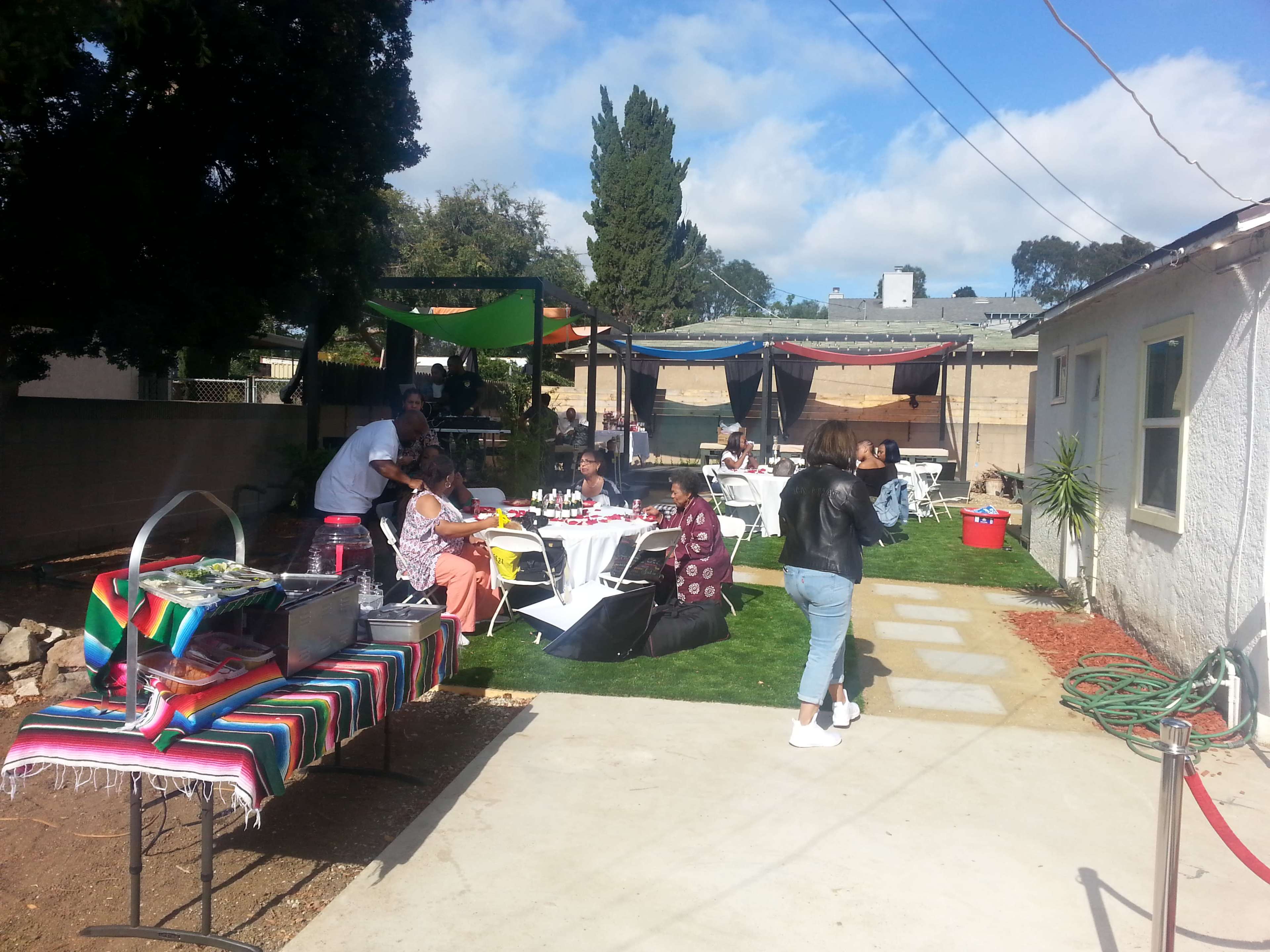 A gathering in a backyard features tables set for dining, a buffet area, and guests engaging in conversation under colorful canopies.