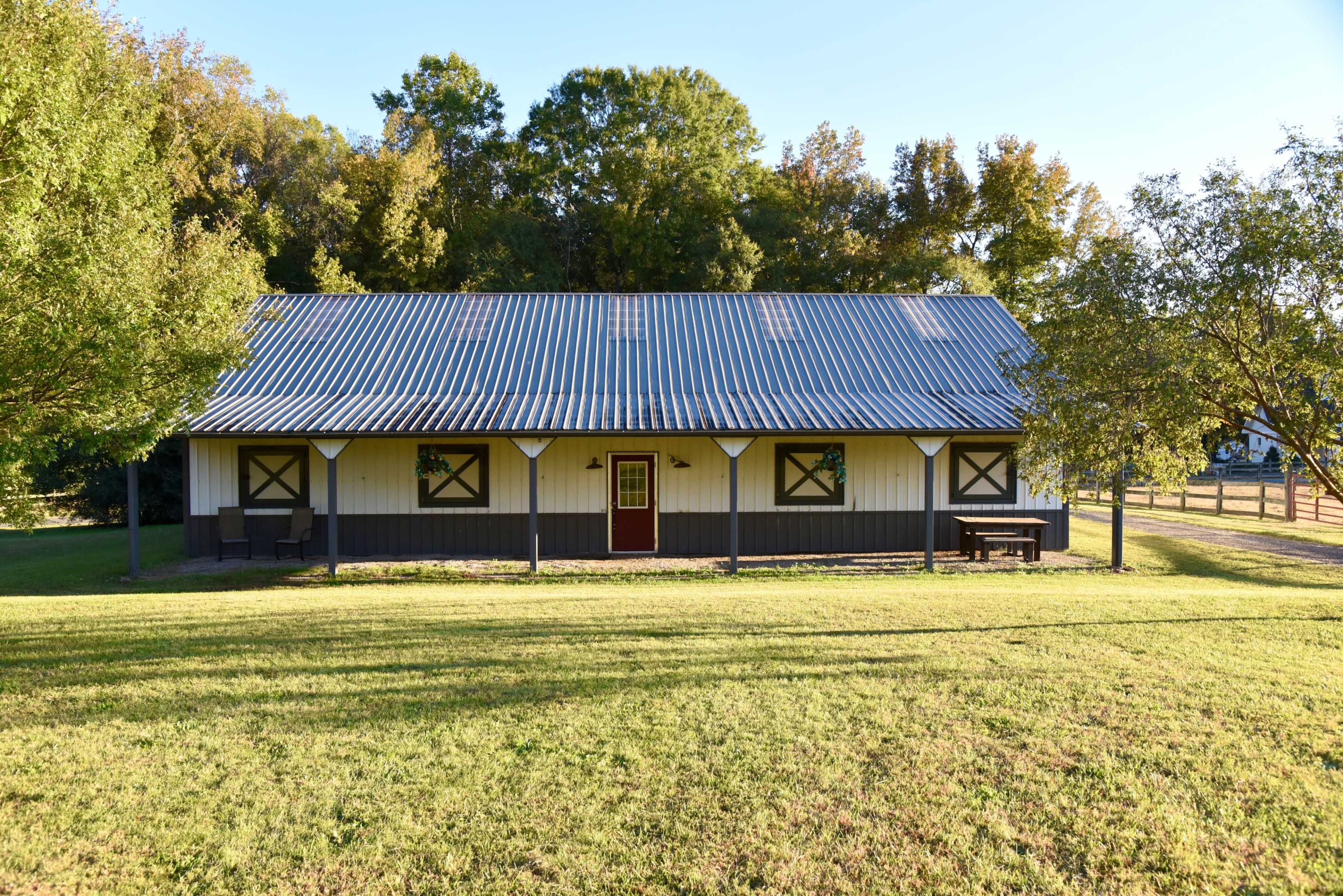 A single-story building with a metal roof and symmetrical windows is situated on a grassy area surrounded by trees.