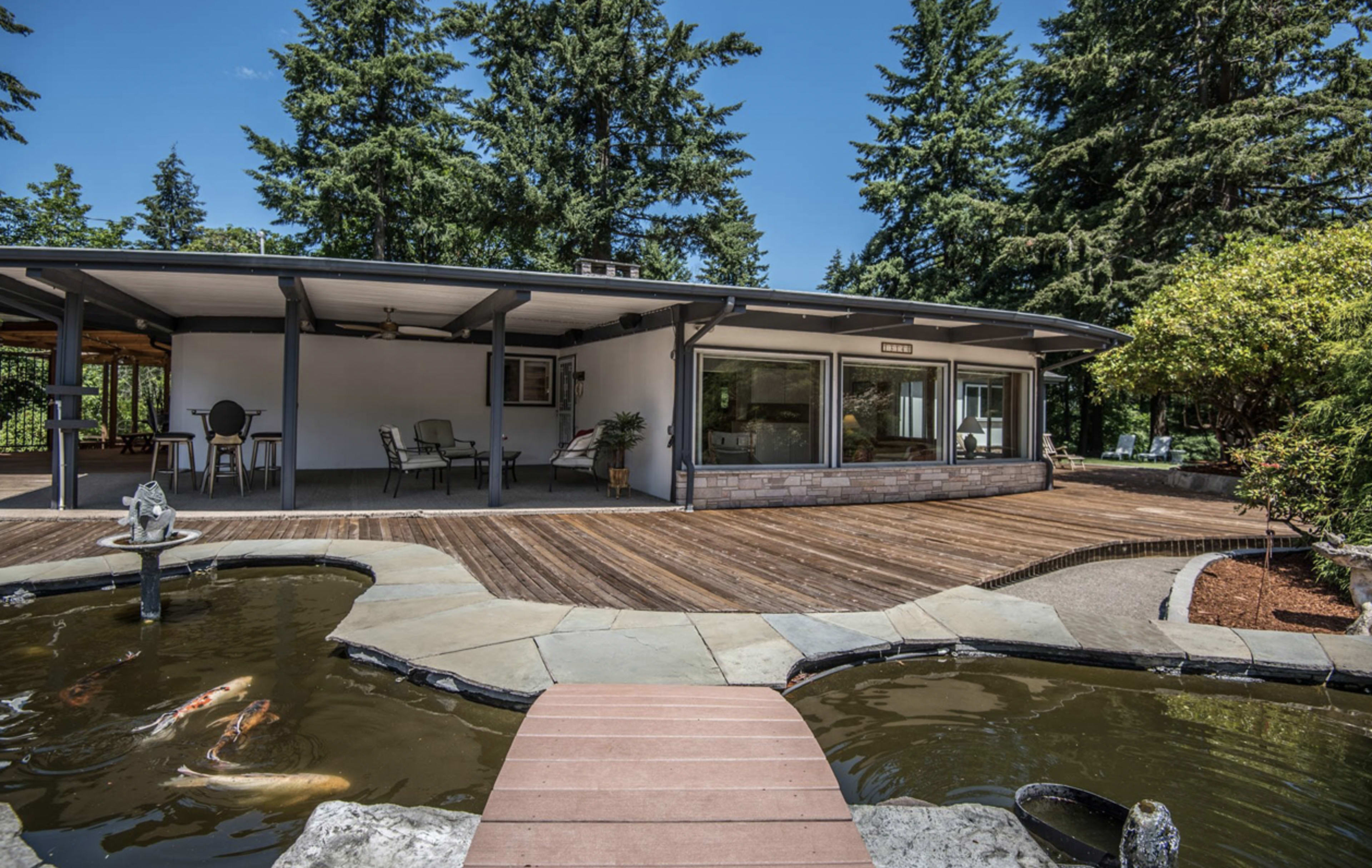 A modern house with a wooden deck, adjacent to a pond featuring koi fish, surrounded by tall trees.